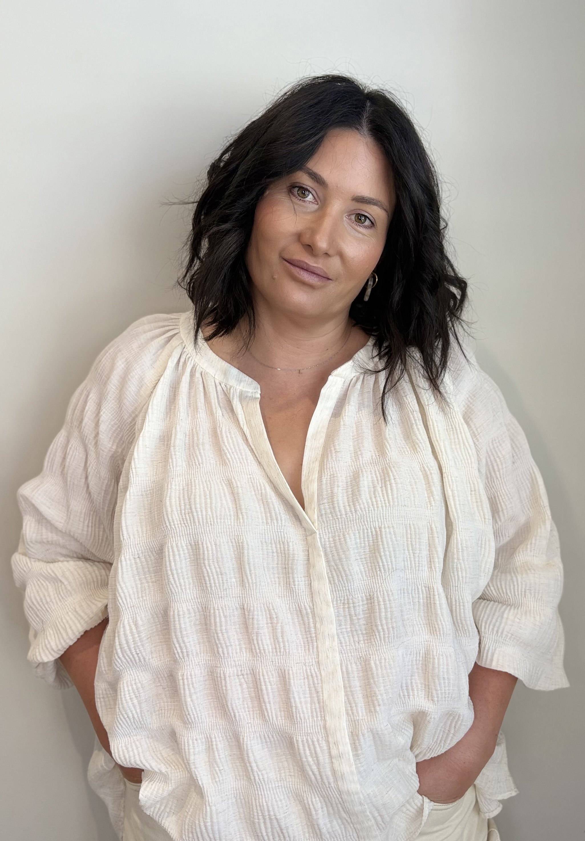 Liz Larner skin specialist, beauty therapist, dermal therapist with shoulder-length dark wavy hair wearing a cream-colored textured blouse standing against a plain light-colored wall. Mount Martha, Mornington Peninsula