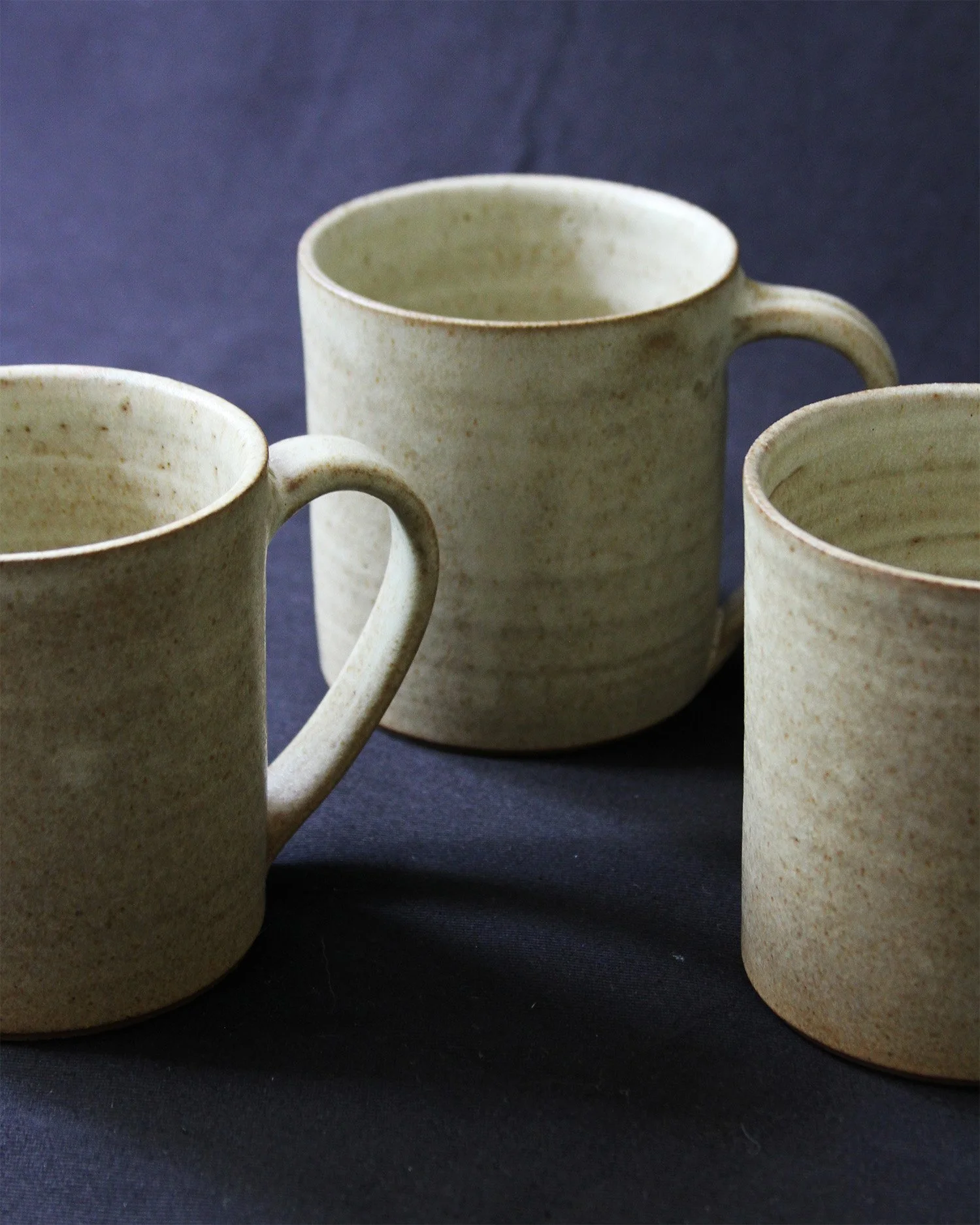 A trio of tea mugs in a sandy speckled glaze on a deep blue background