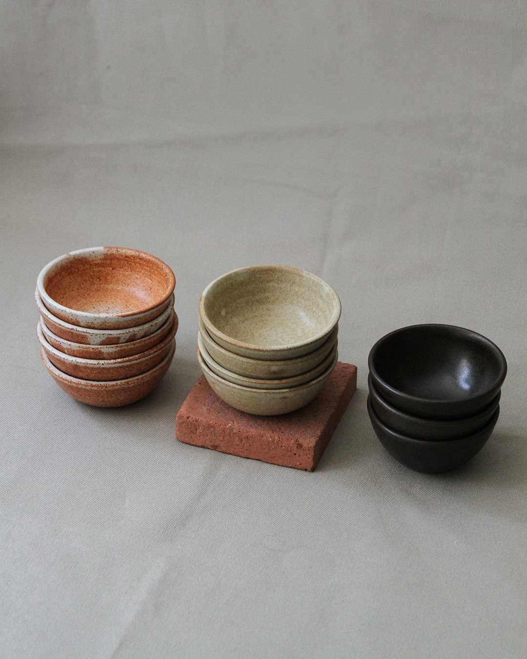 Three stacks of little bowls against a light green background. On the left are rusty speckled bowls, in the middle bowls with a sandy glaze, and on the right bowls in a rich black-brown glaze.