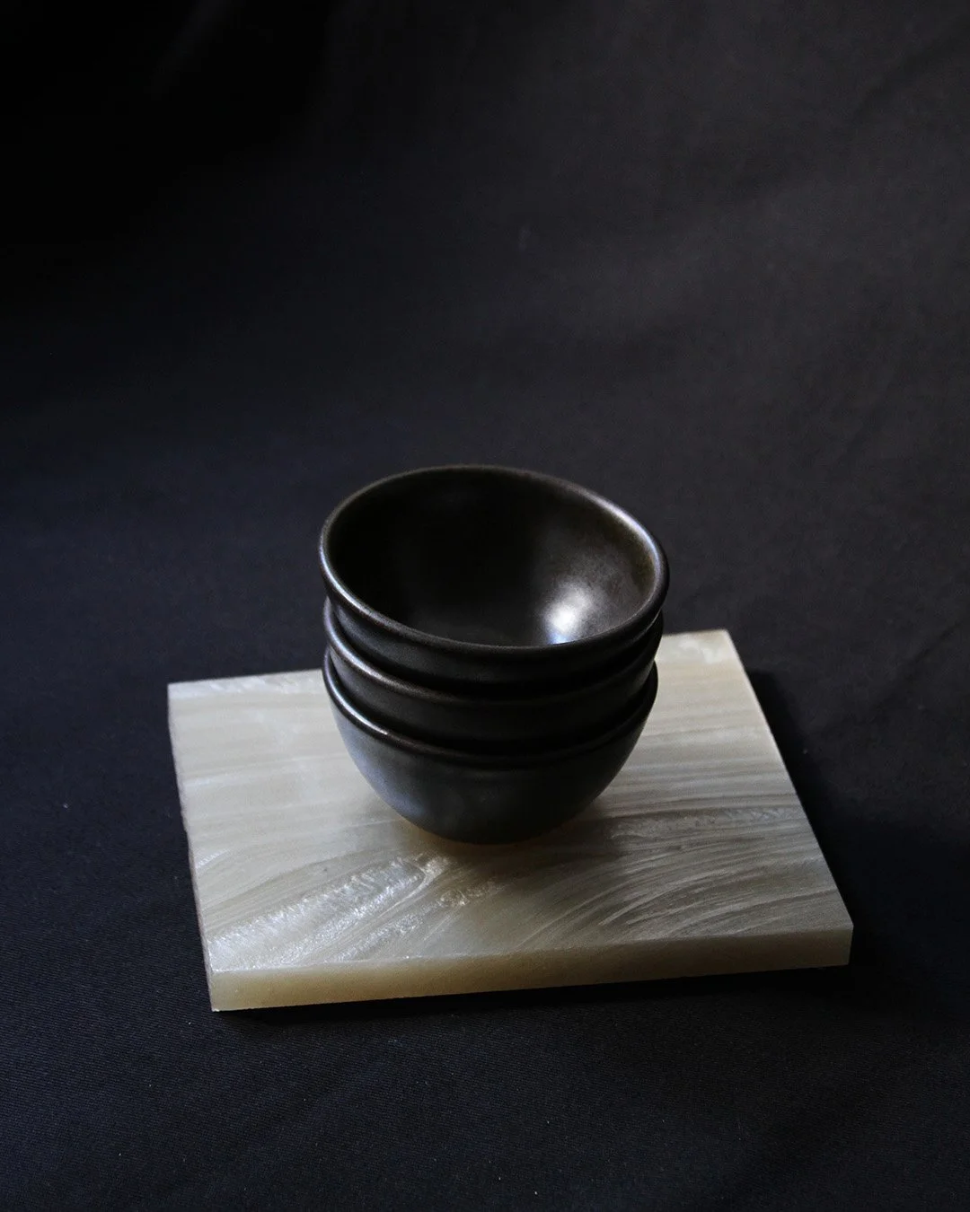 A stack of little bowls in a rich black-brown glaze, placed on a pearlescent plinth with an organic ridged texture.