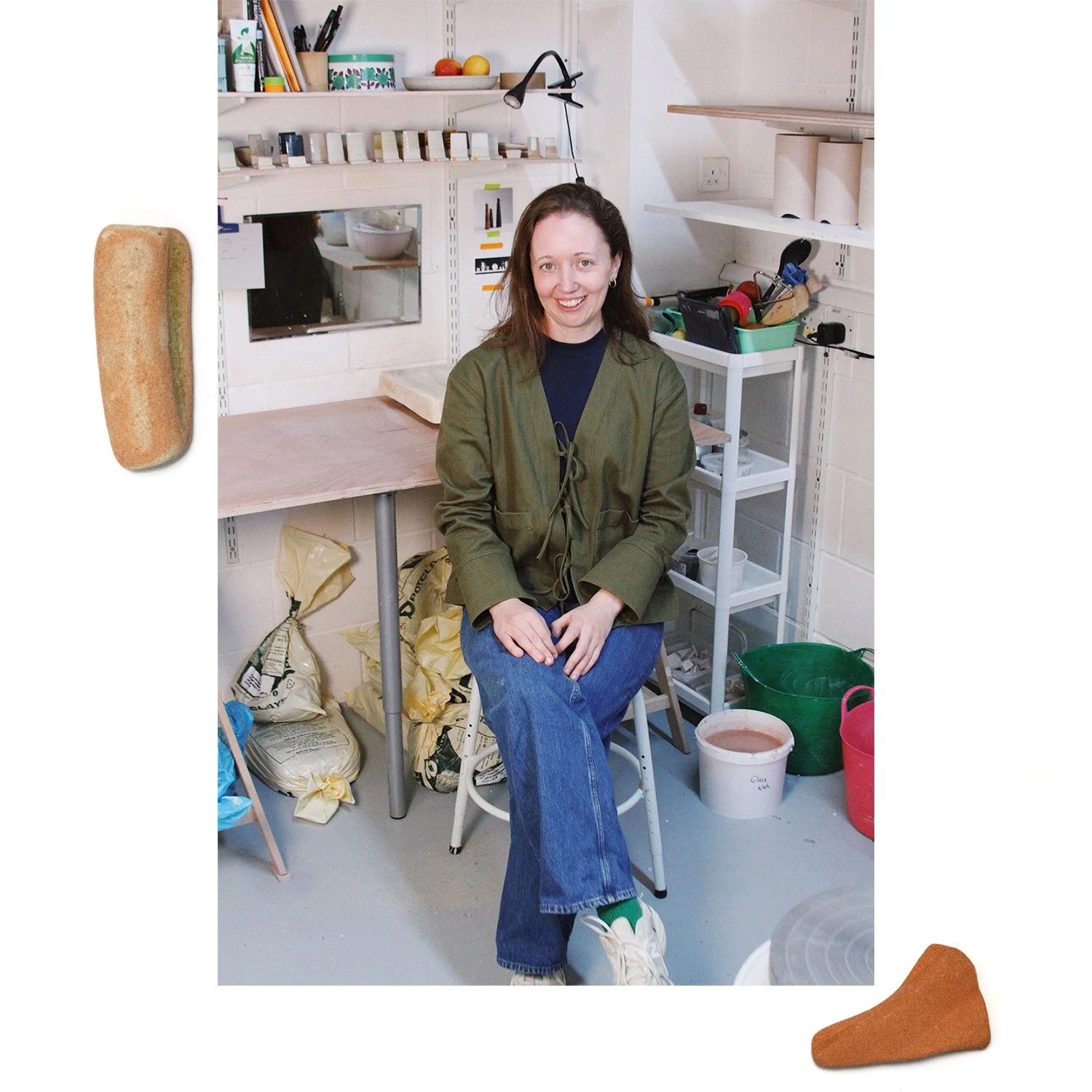 A woman sitting on a stool in a workshop or studio, smiling at the camera. She is wearing a green jacket, blue jeans, and white sneakers. The background includes shelves with various supplies, a small mirror, and bags of materials on the floor. There are also two rocks or stones, one on the left side and one on the bottom right corner of the image.