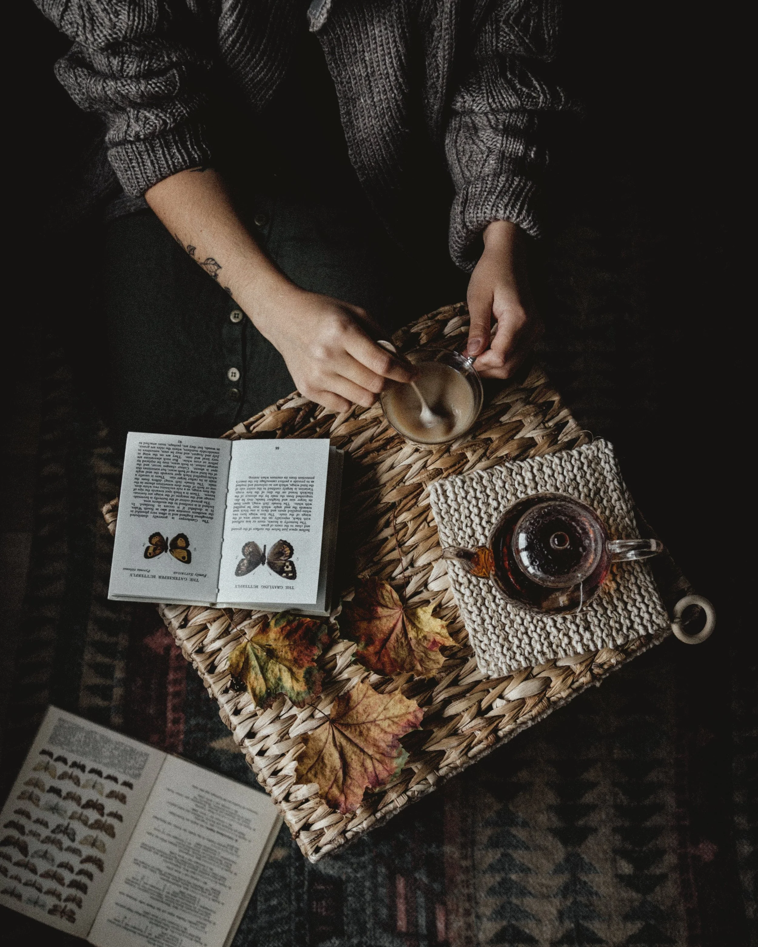 Persona sentada en una alfombra de la sala, con un libro abierto y varias hojas de libro, tomando una taza con una bebida caliente. Hay una tetera y una taza con infusión, hojas de otoño en la mesa de mimbre, y un tapete con patrón geométrico debajo.