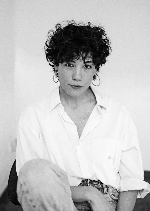 Retrato en blanco y negro de una mujer con cabello rizado y curto, con camiseta blanca y pantalones vaqueros, mirando a la cámara