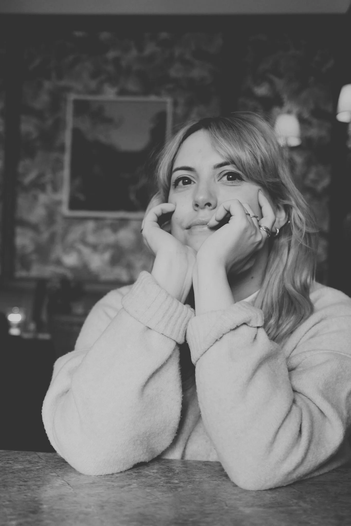 A black-and-white selfie of a woman with medium-length wavy hair, wearing a sweater and small earrings, with a poster of structures behind her on the wall.