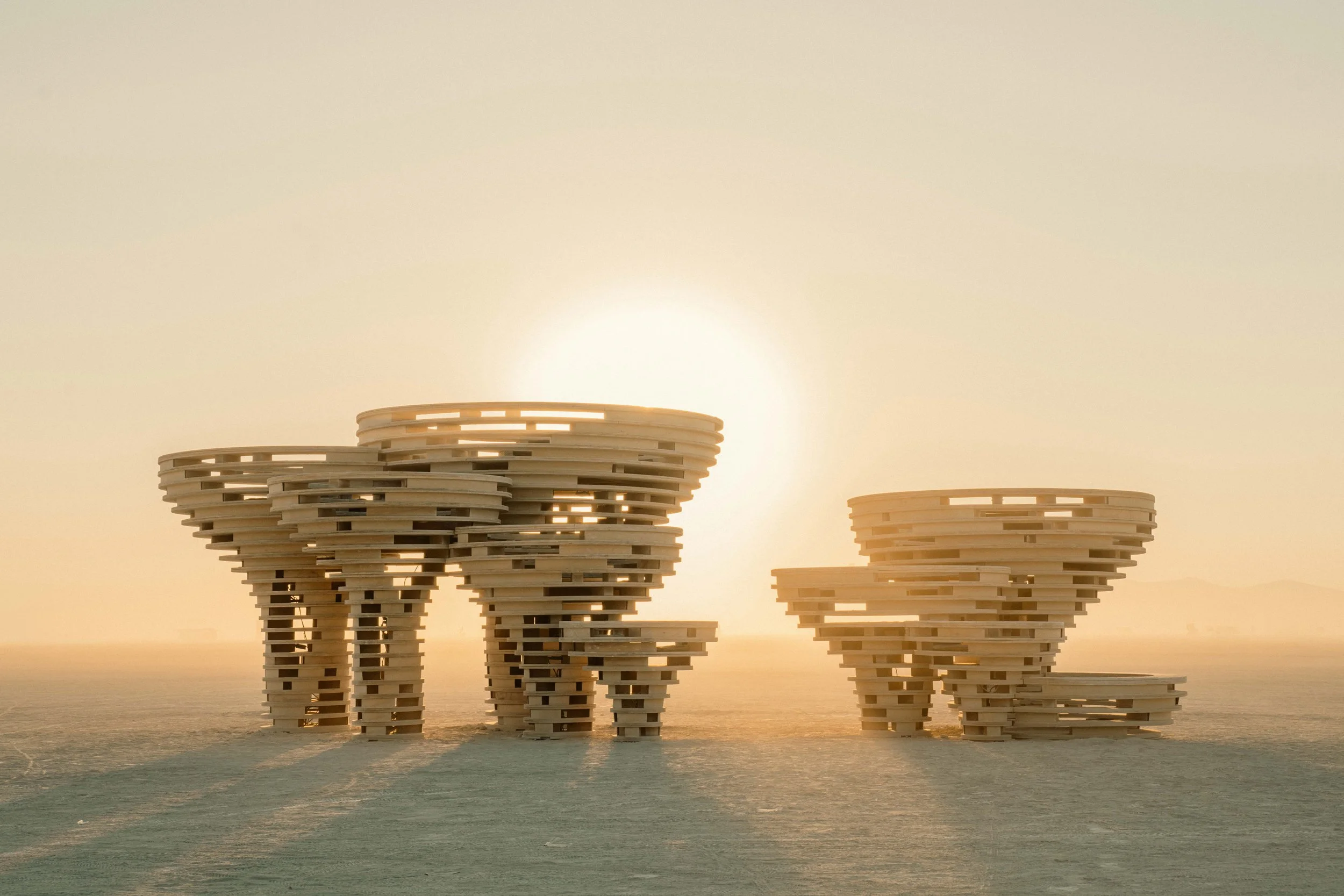 Architectural structures made of stacked wooden planks in the shape of abstract towers, set against a sunset or sunrise in a desert-like landscape.