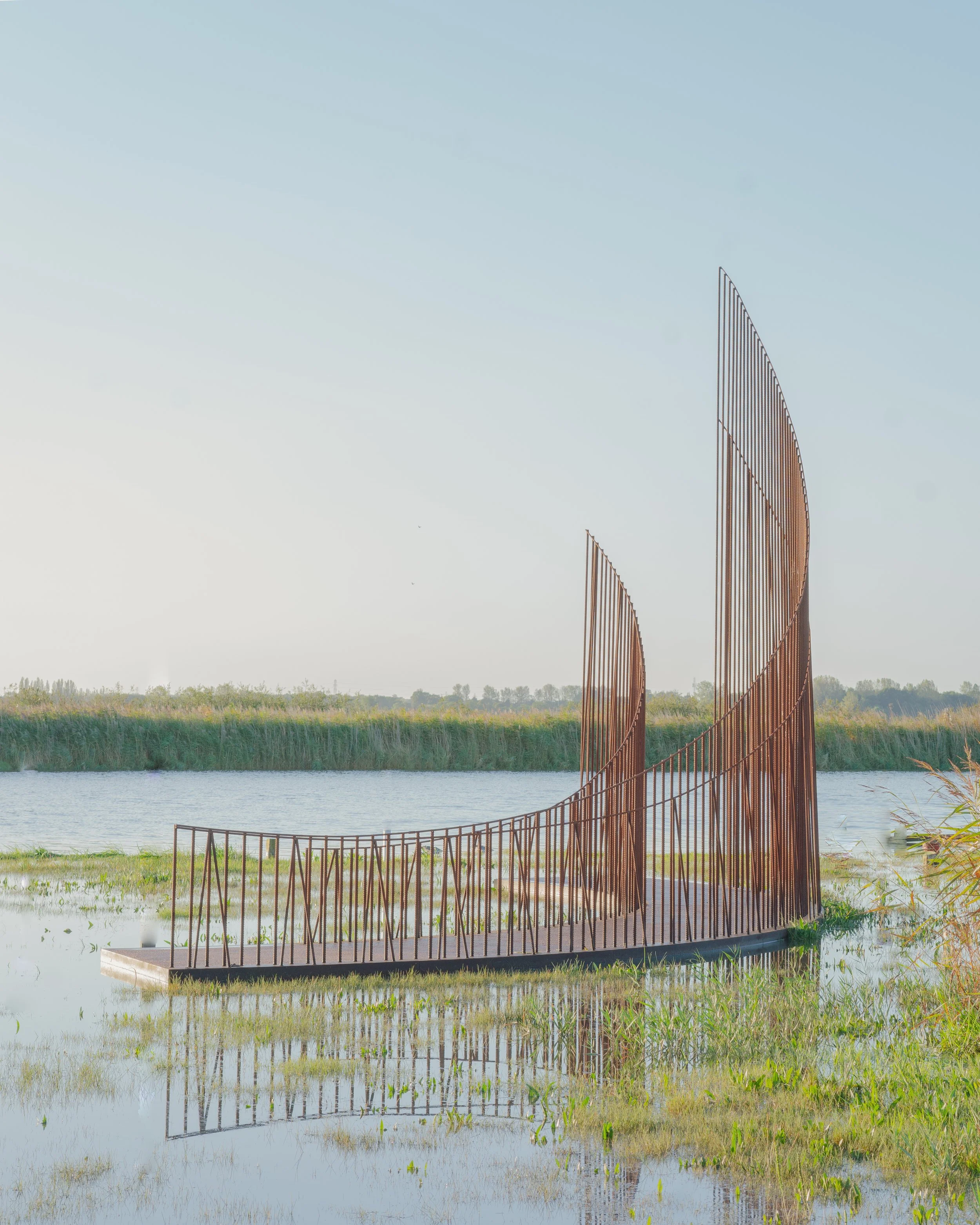 Sculptural art installation with rust-colored metal rods forming an abstract structure near a body of water, surrounded by marshland and tall grasses, under a clear sky.