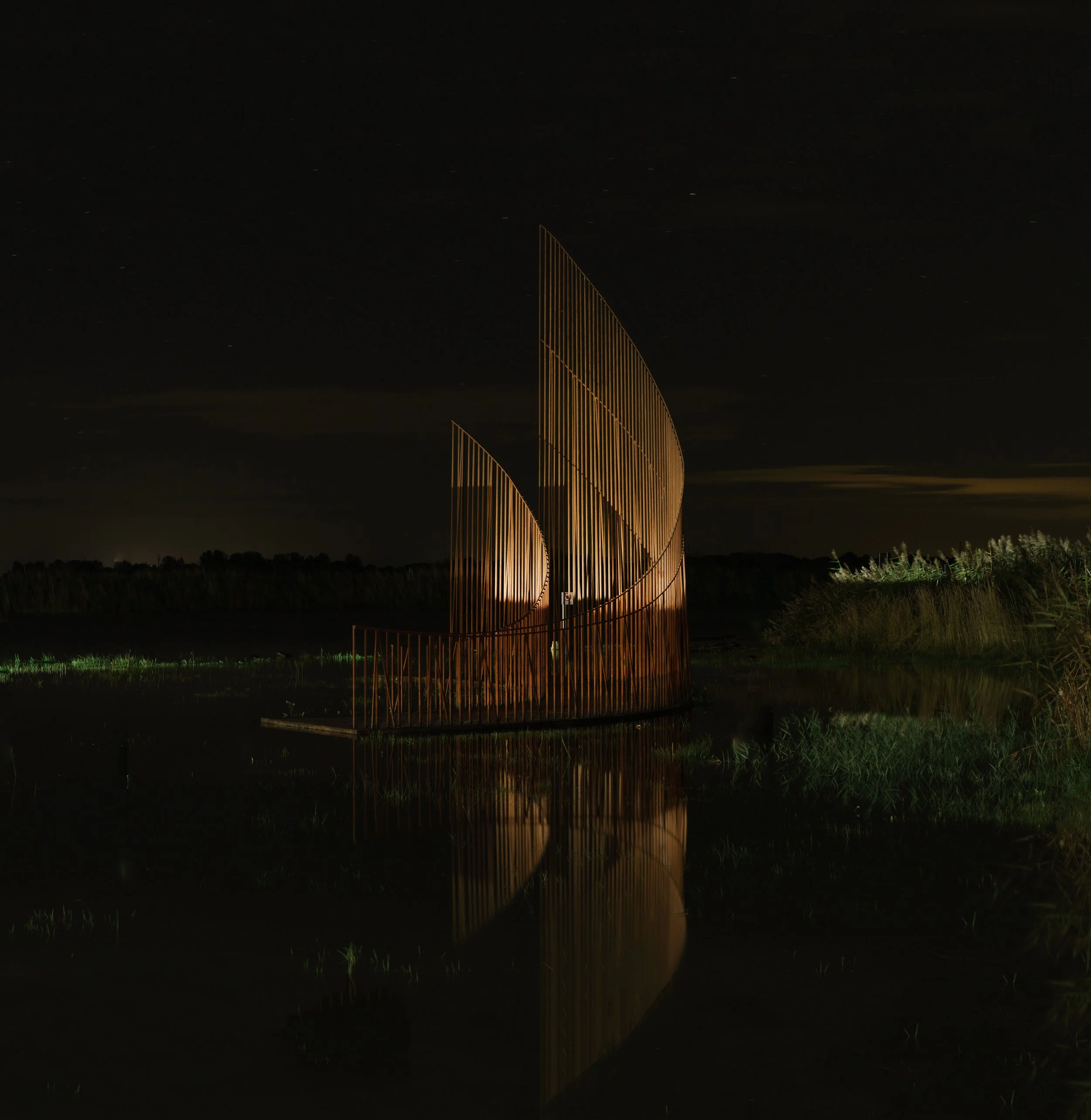 Night view of a modern outdoor sculpture with vertical golden bars forming curved shapes, reflected in water, against a dark sky with distant stars.