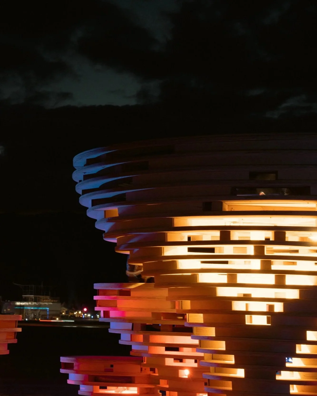 Nighttime view of a modern circular sculpture made of stacked wooden slats with internal orange lighting, under dark cloudy sky.