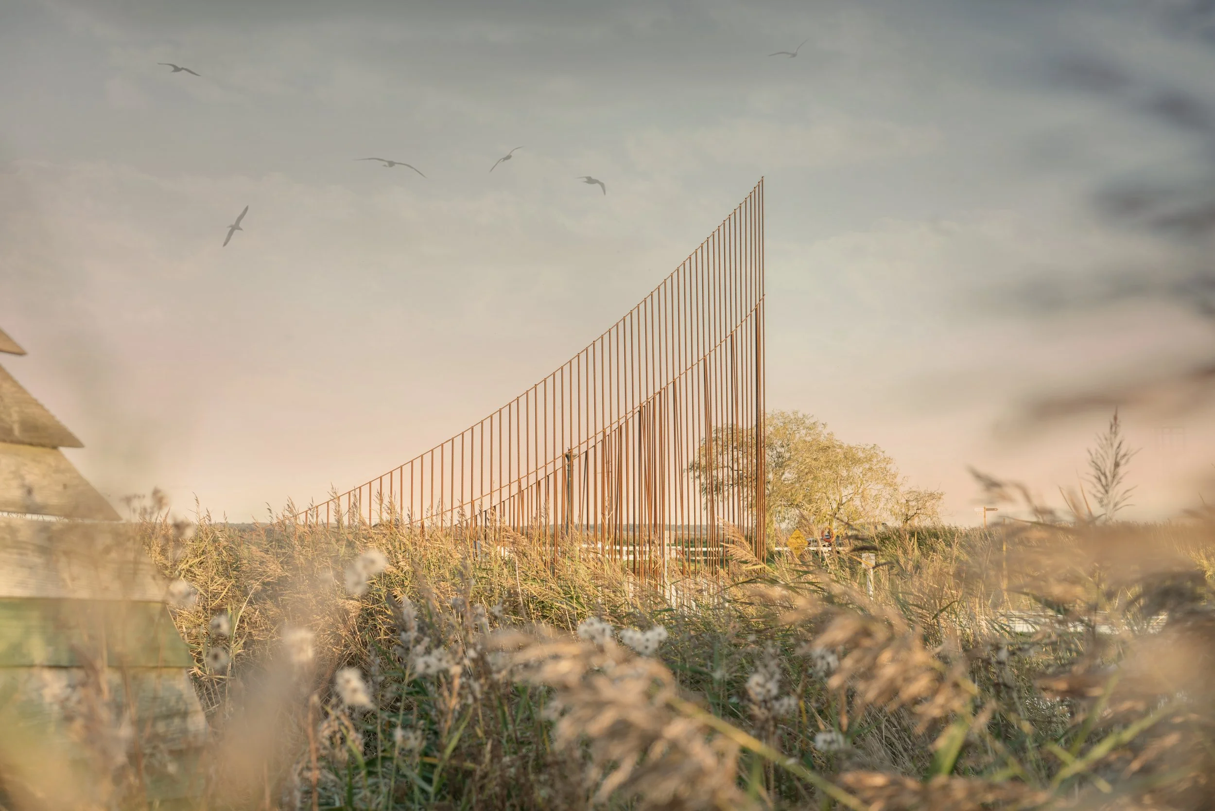 A tall, modern metal fence structure in an open field with grass and trees, under a sky with cirrus clouds and birds flying.