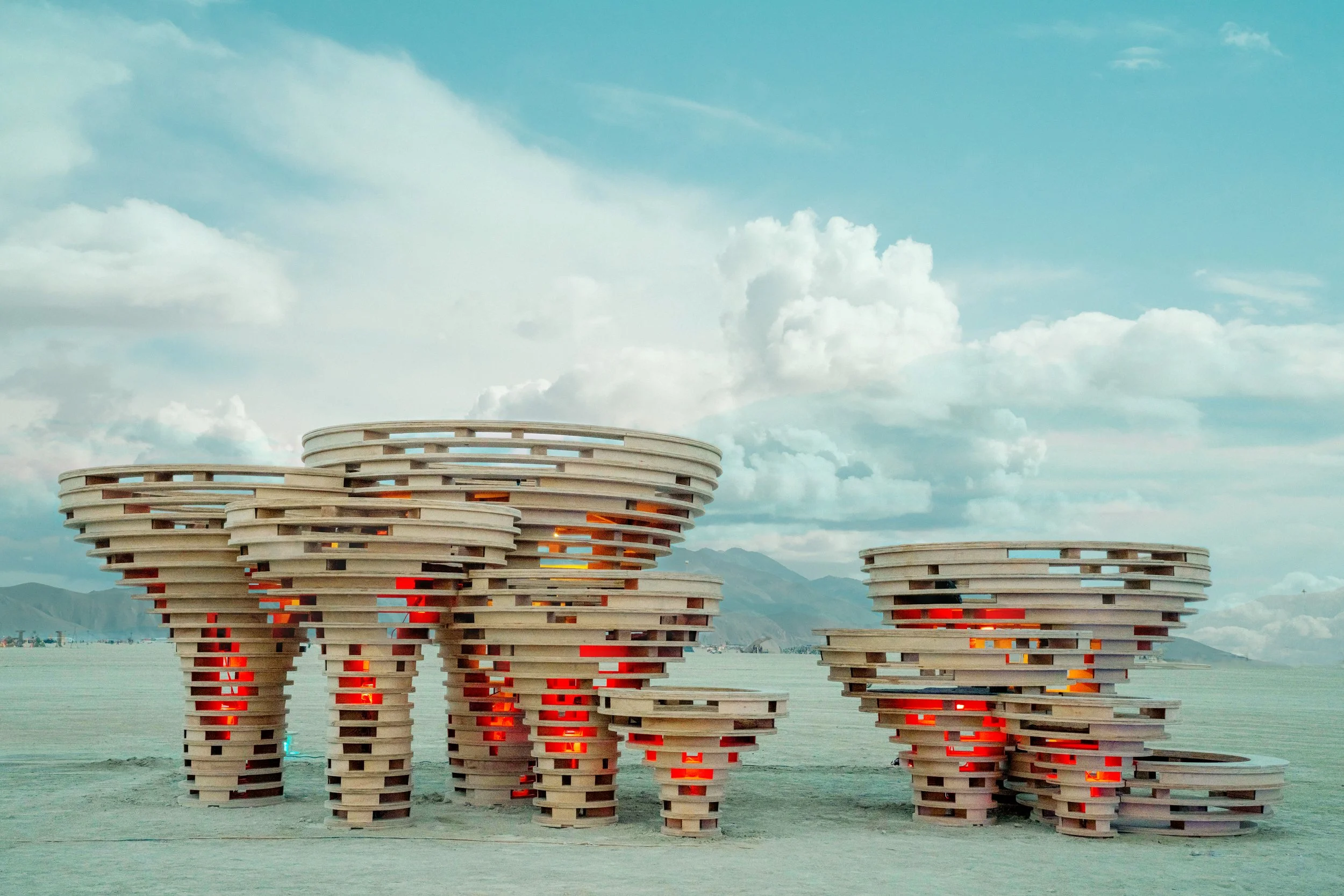 Contemporary art installation with stacked circular wooden structures with red and orange lighting inside, set in a vast open landscape with mountains and partly cloudy sky in the background.