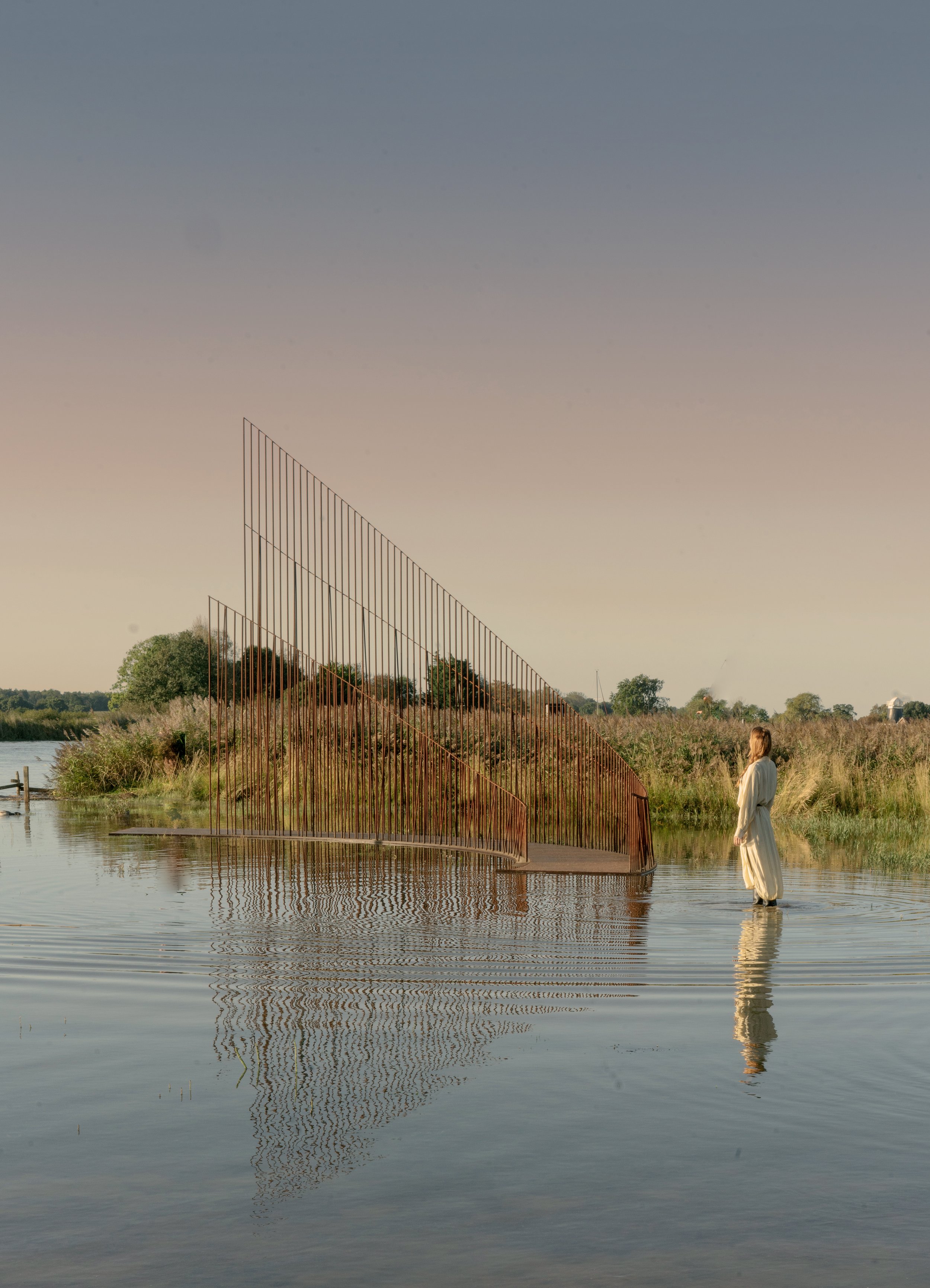 A person in a long white dress standing in shallow water near a modern sculpture made of rust-colored metal bars, with a natural landscape and cloudy sky in the background.