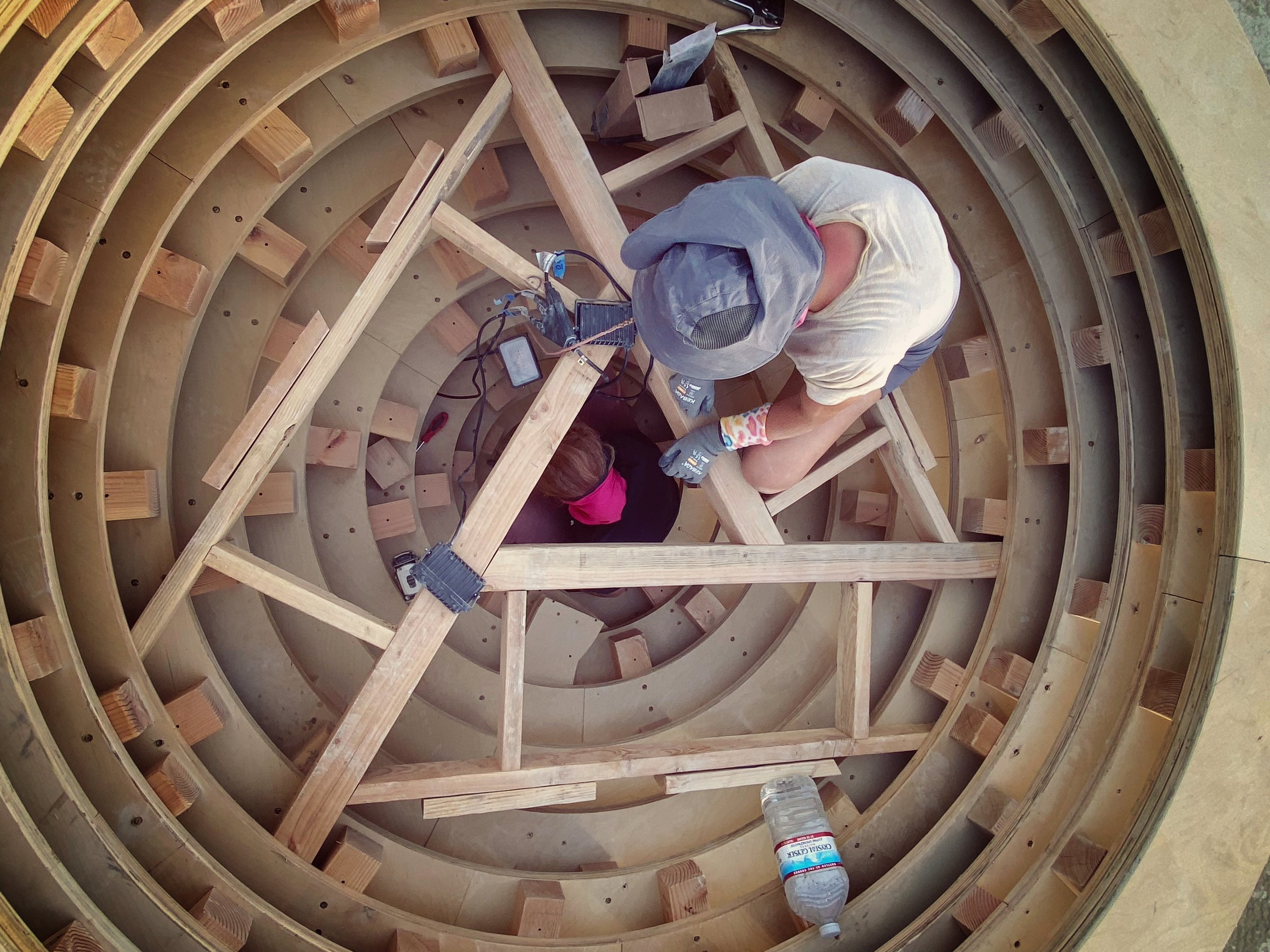Two construction workers are building a wooden spiral staircase, viewed from above.