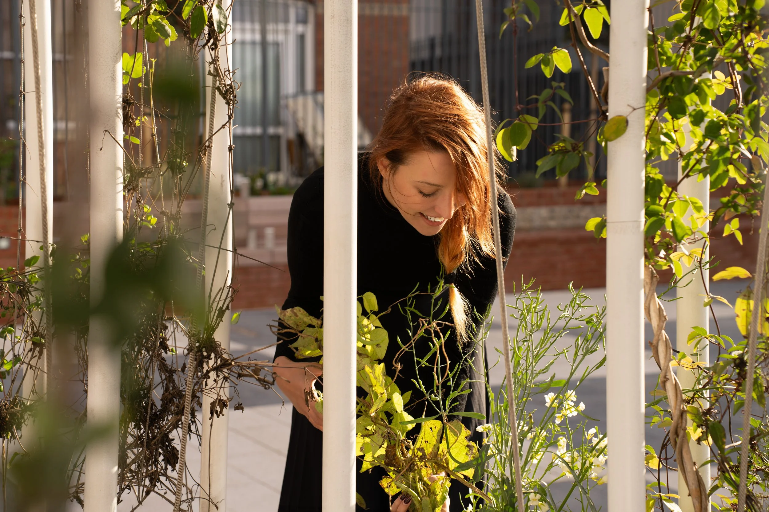 A woman with red hair, smiling, tending to plants on a rooftop garden, surrounded by white metal posts and greenery.