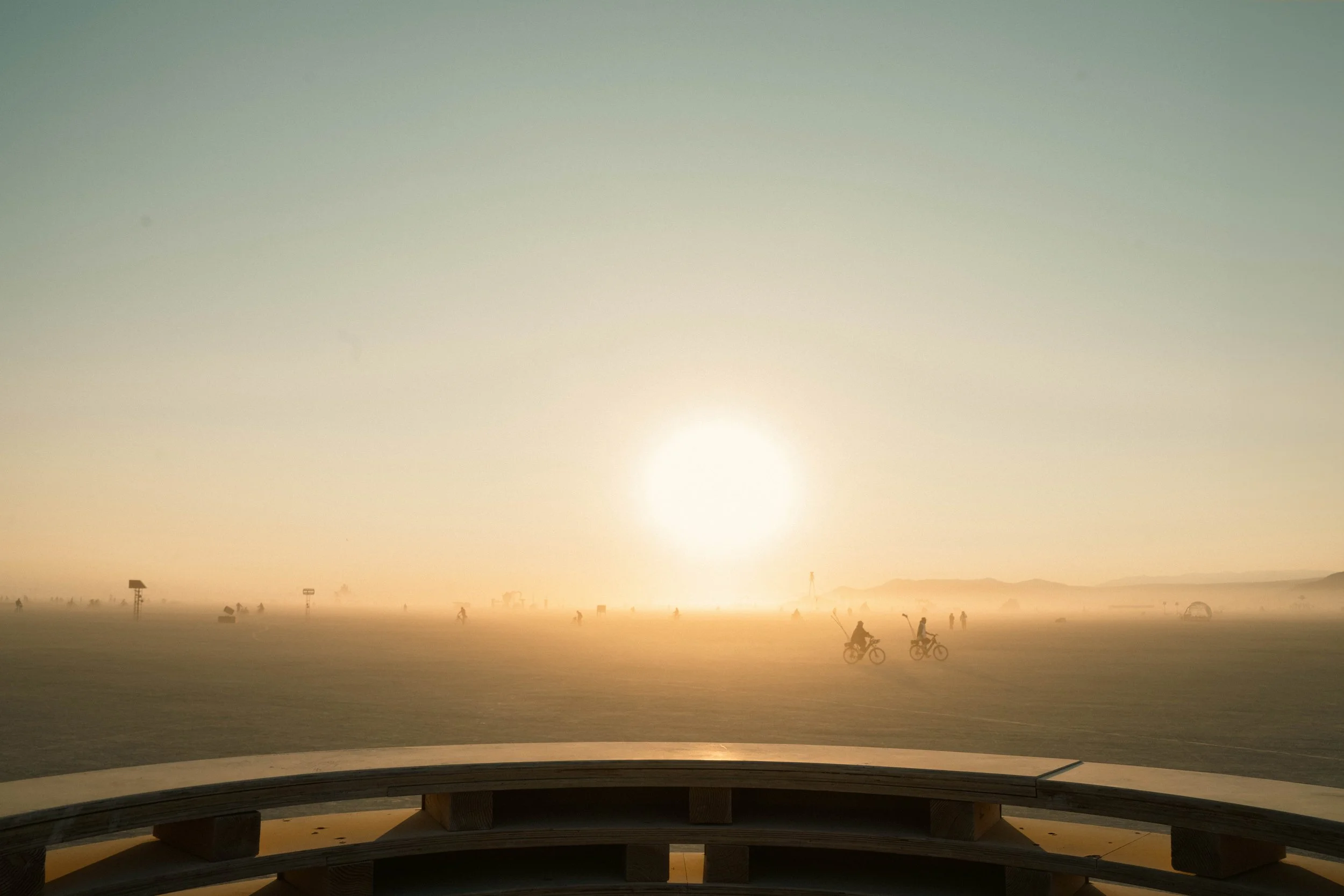 A vast flat landscape with a group of people riding bicycles, silhouetted against a foggy horizon with a large, bright sun and mountains in the distance, viewed from a wooden structure.