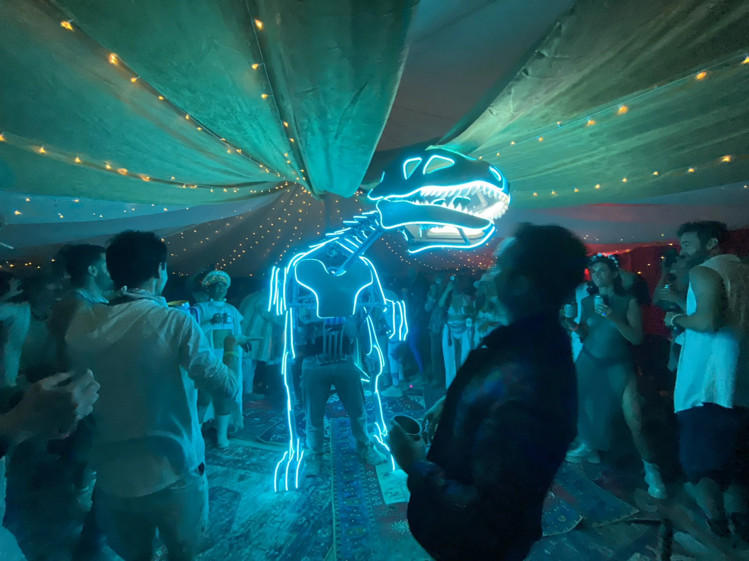 People dancing and socializing at a party with a neon dinosaur sculpture illuminated in blue, under a ceiling decorated with string lights.