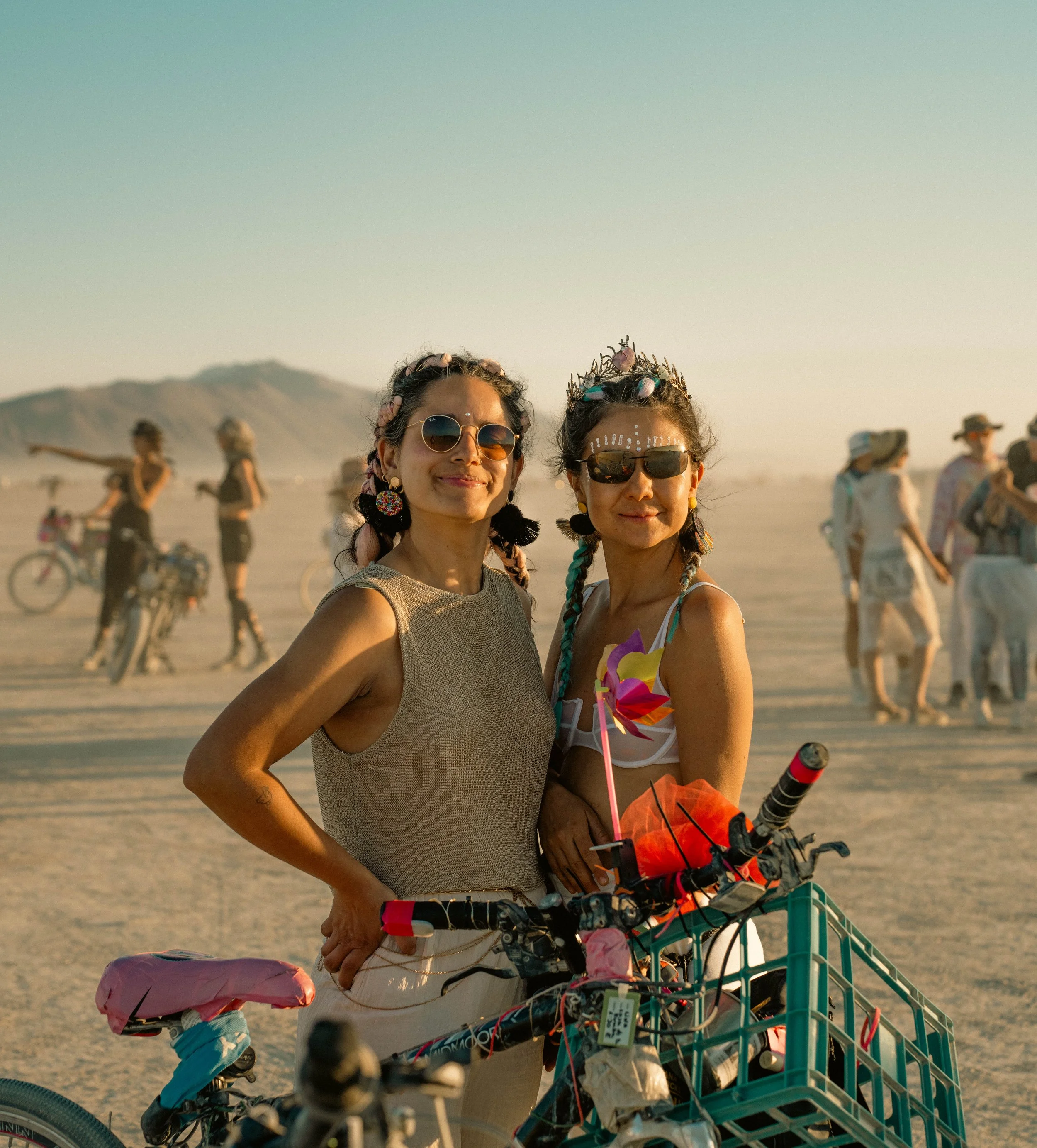 Two women with colorful hair and accessories posing with their bikes at a desert festival.