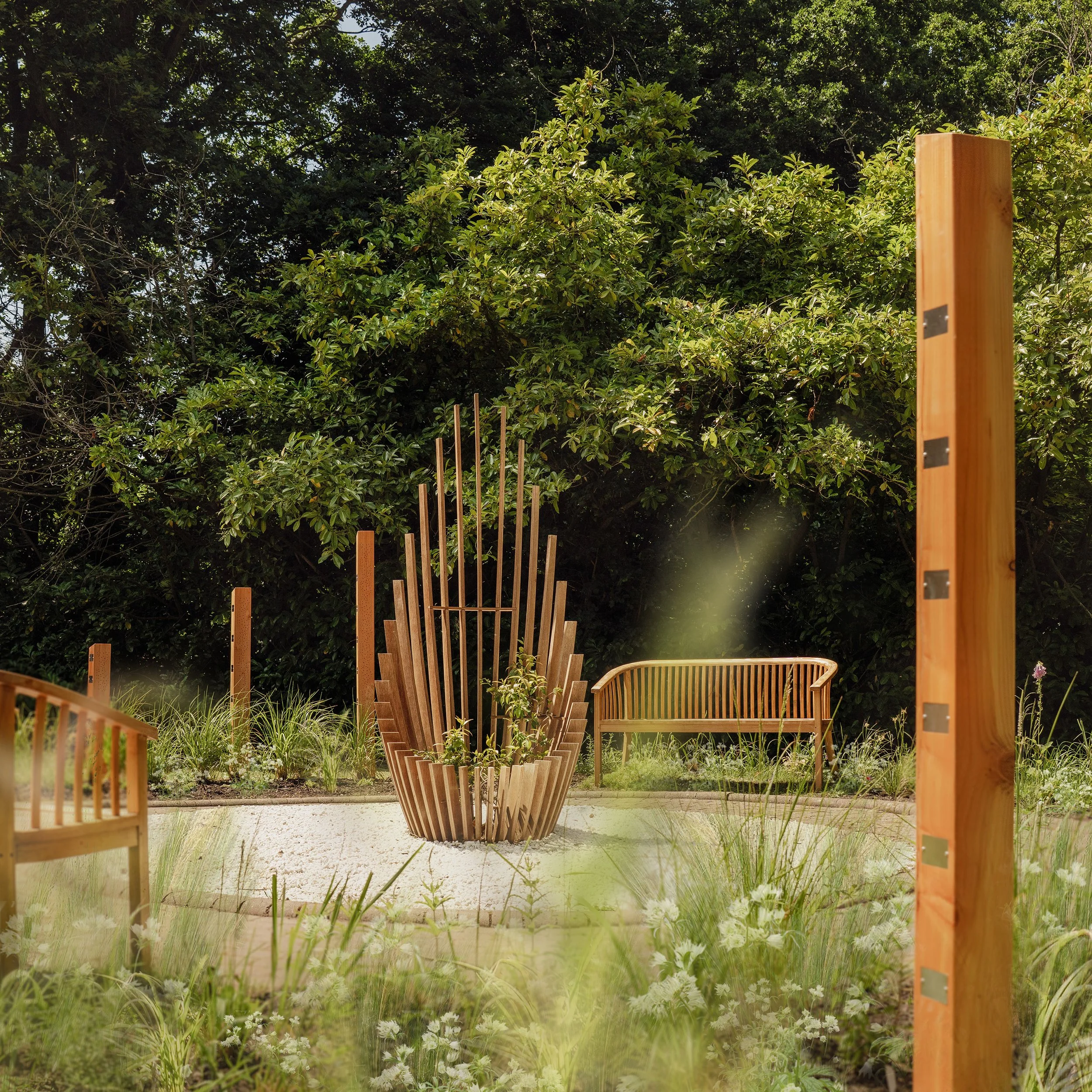 A landscaped outdoor space with a modern wooden sculpture, a wooden bench, and wooden posts among lush greenery and flowers.