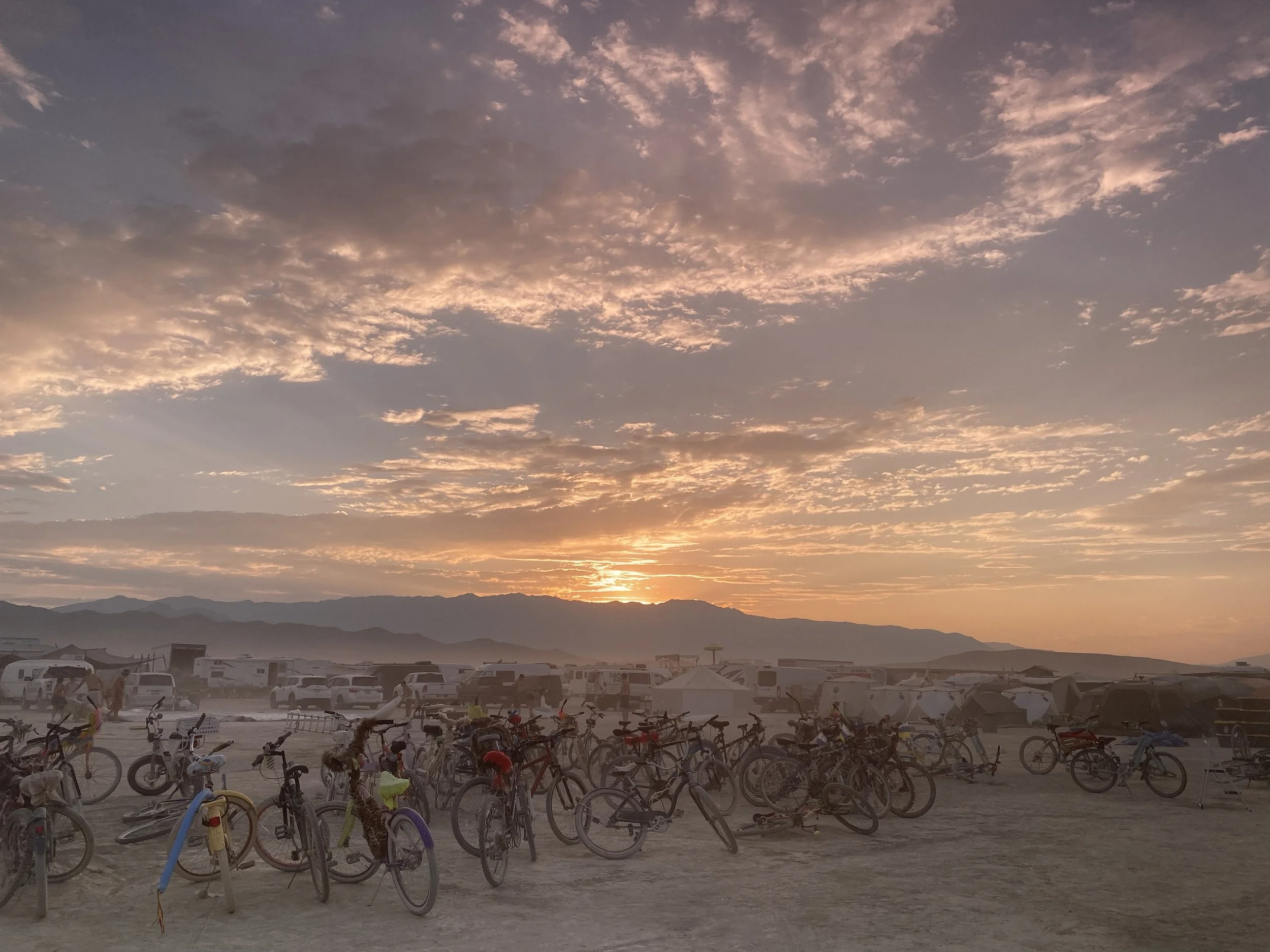 Bicycles parked on a dusty desert terrain with tents and vehicles in the background during sunset, mountains are visible in the distance under a colorful sky with scattered clouds.