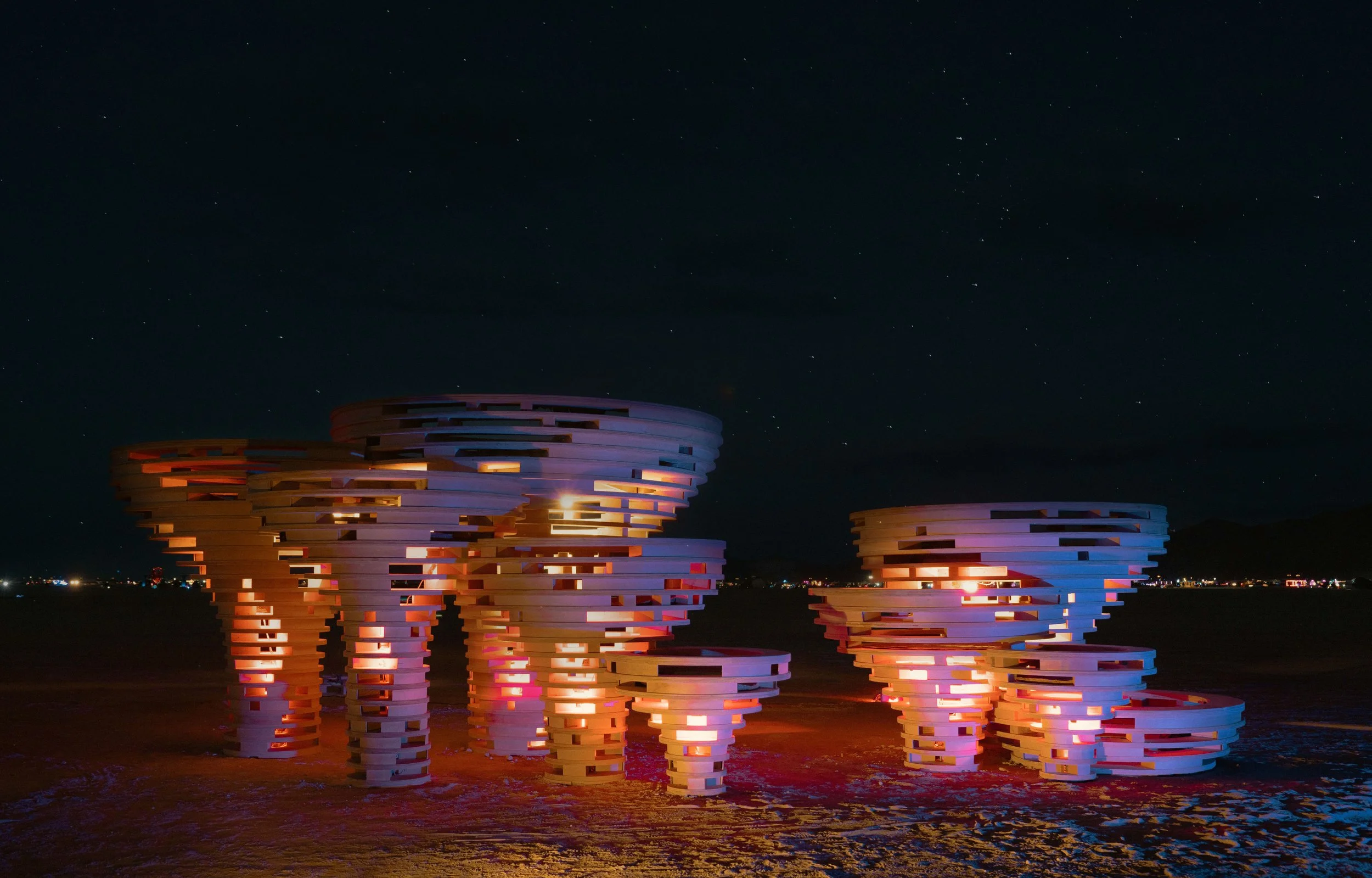Nighttime view of futuristic, sculptural structures resembling stacked horizontal bands illuminated with colored lights, set against a dark sky filled with stars and a distant city skyline.