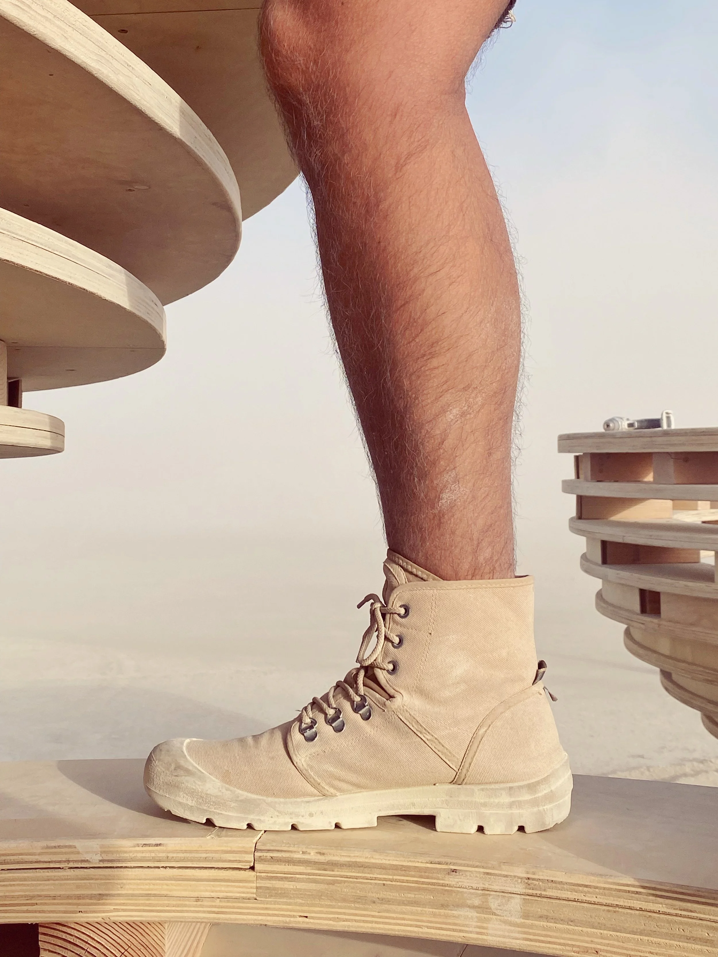 A person's leg wearing a beige high-top boot standing on a wooden surface with a curved design, with another wooden object and an open sky in the background.