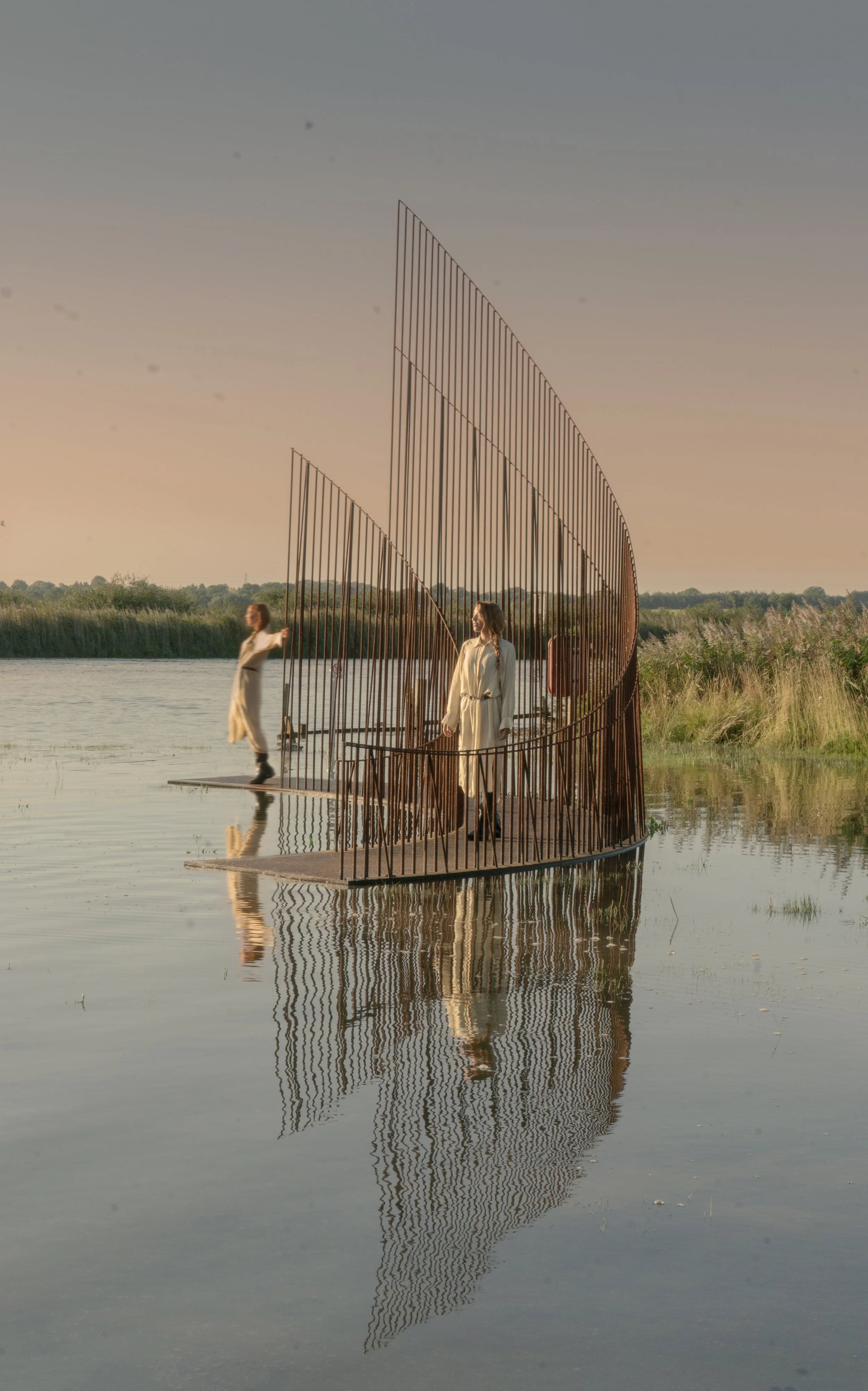 Three women in beige coats standing and walking on a modern metal installation on the water during sunset, with reflections and a natural landscape in the background.