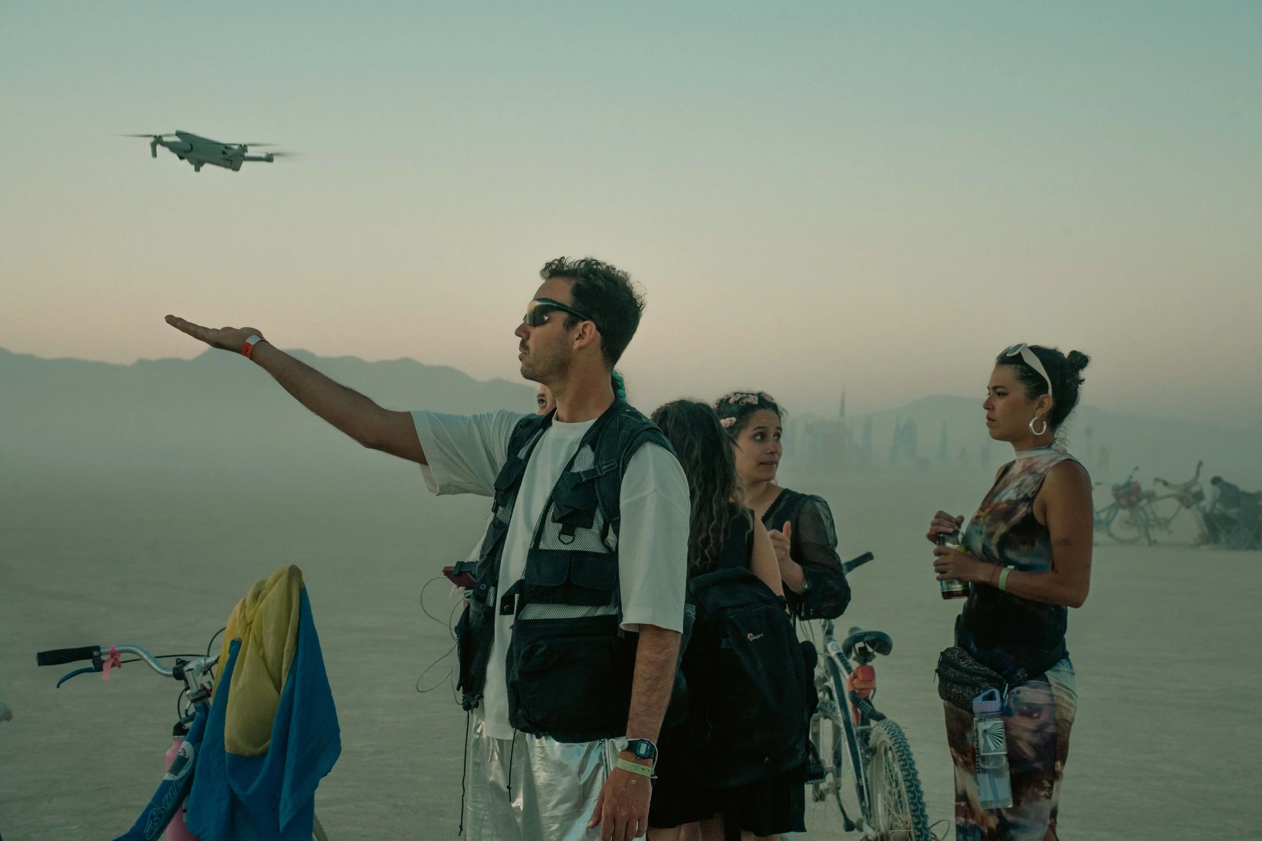 A group of four people standing on a salt flat with bicycles and a drone flying in the background. The man in front is extending his arm, pointing at the drone, wearing sunglasses, a white t-shirt, and a backpack. The women are gathered around, with 