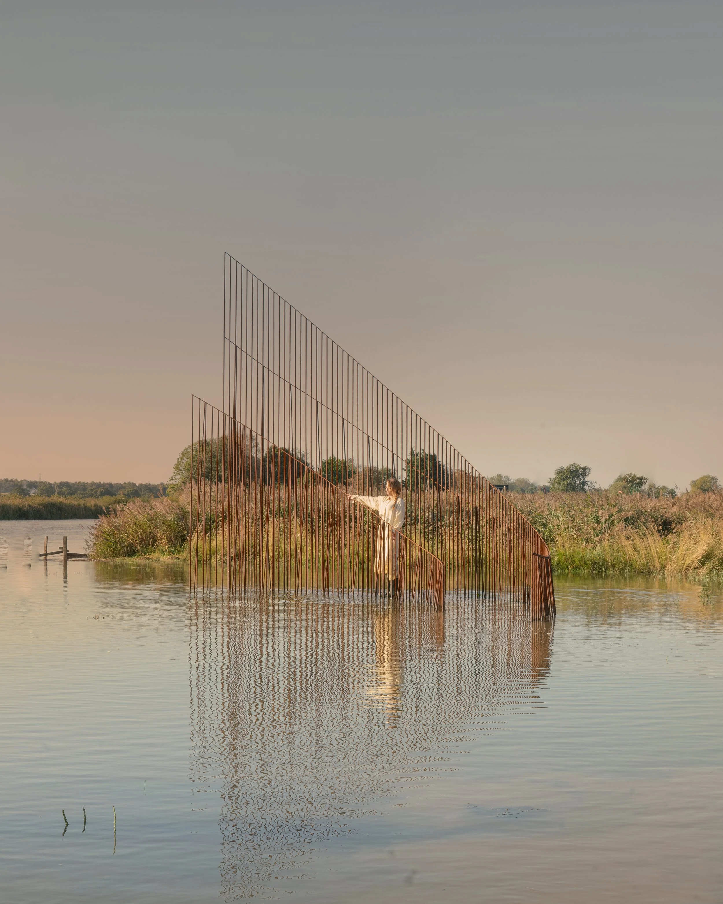 A person standing in water near a tall, abstract metal sculpture with slanted rods forming geometric shapes, surrounded by nature.