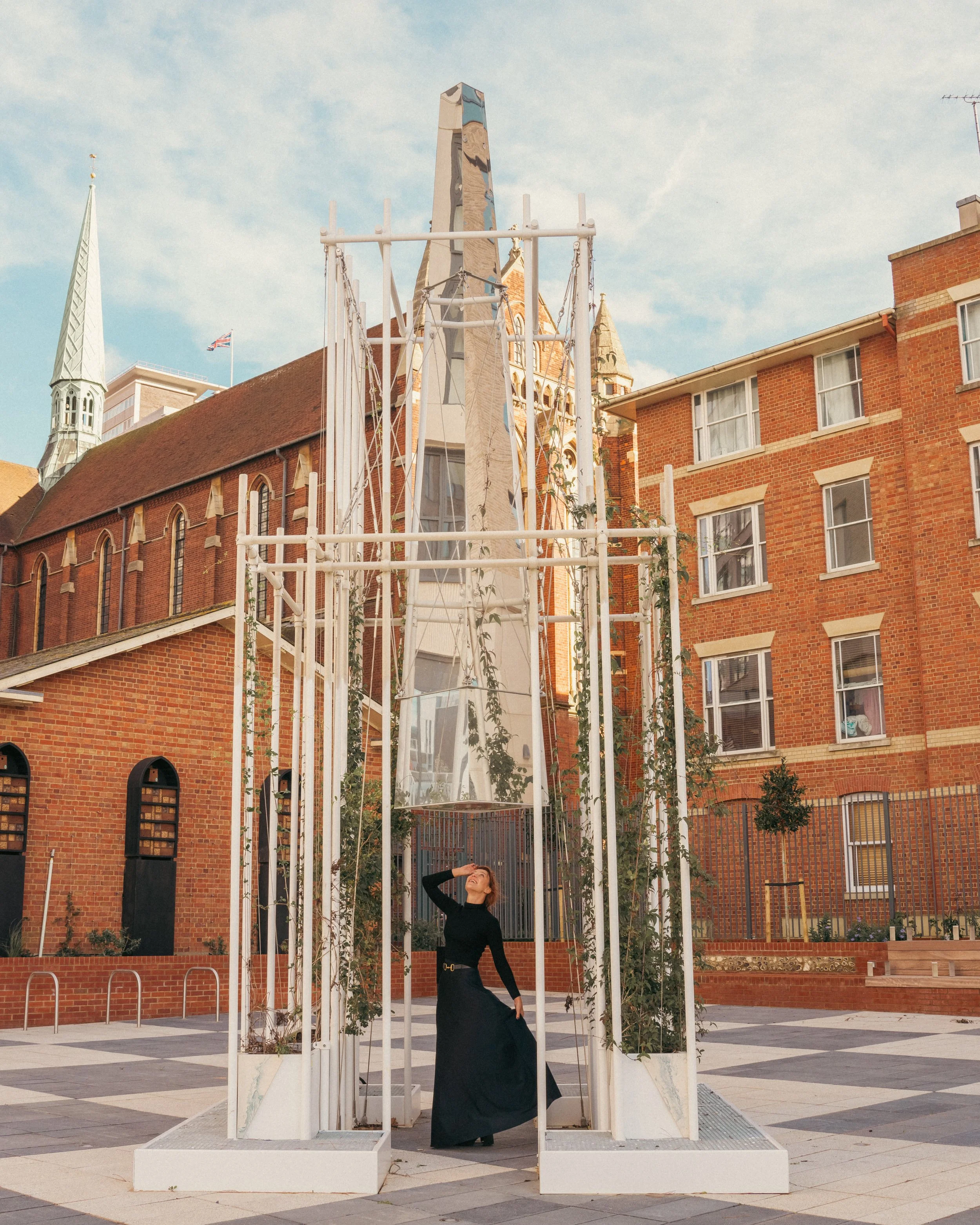 Woman in black dress standing inside a modern white structure with vertical bars and climbing plants, in an outdoor urban setting with brick buildings in the background.