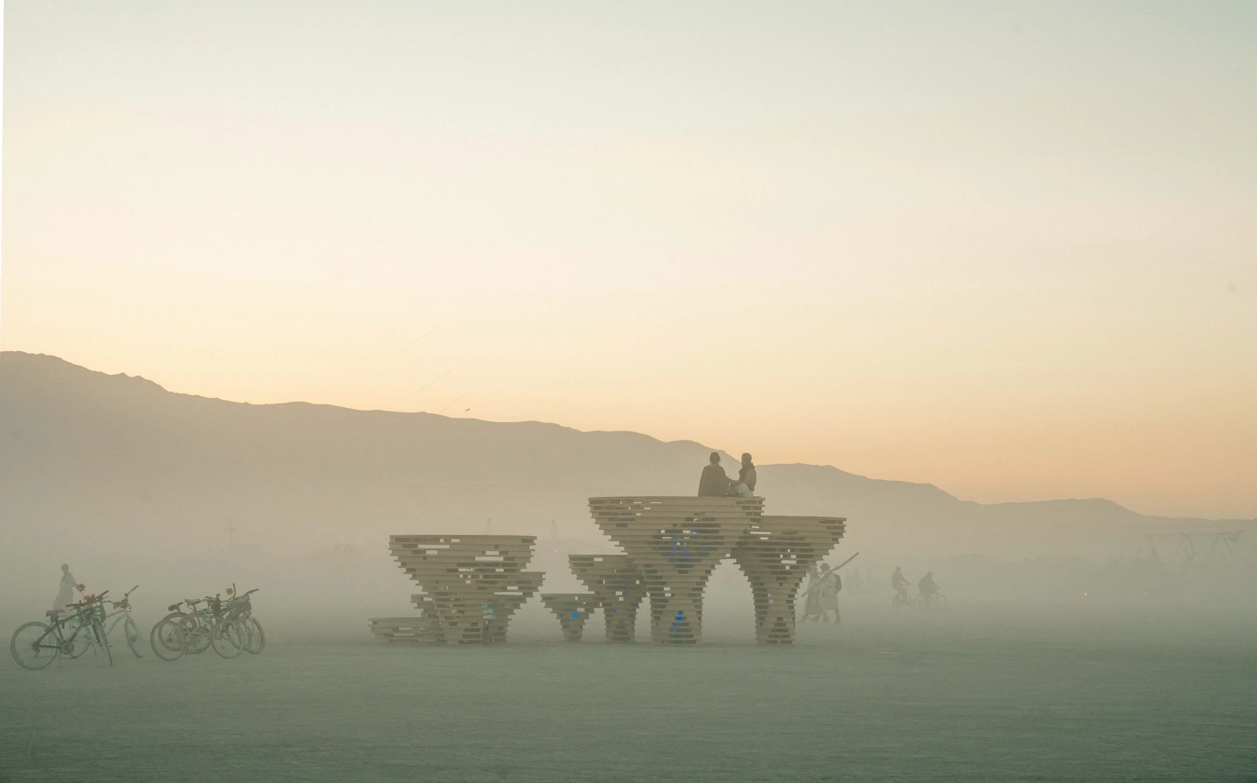 Silhouettes of people sitting on a modern, elevated bench structure in a foggy outdoor setting at dawn or dusk, with bicycles nearby and mountains in the background.