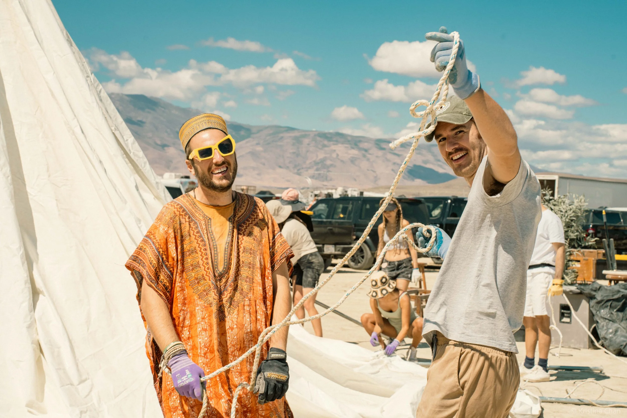 People working together outdoors on a sunny day, setting up a large white tent in a desert landscape with mountains in the background