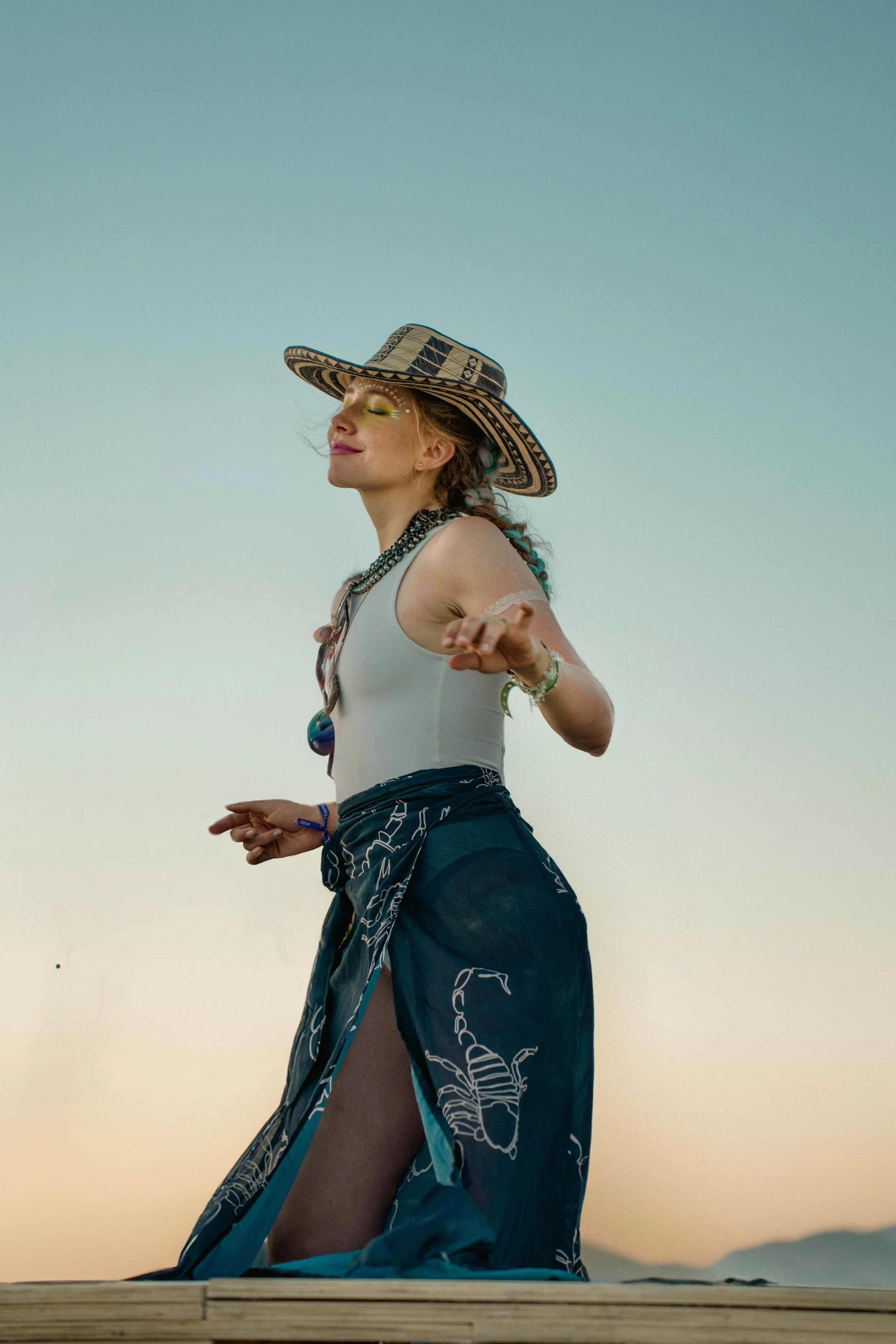 A woman with a large wide-brimmed hat, necklaces, and a sheer skirt with lobster print, standing outdoors at sunset with closed eyes and a peaceful expression.