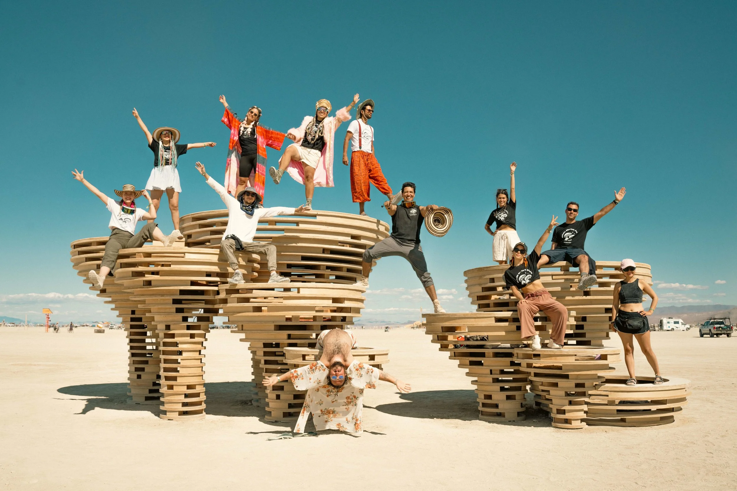 A group of people posing on a large wooden art installation shaped like the word 'LOVE' in a desert setting. Some are sitting, some standing, with a clear blue sky in the background.