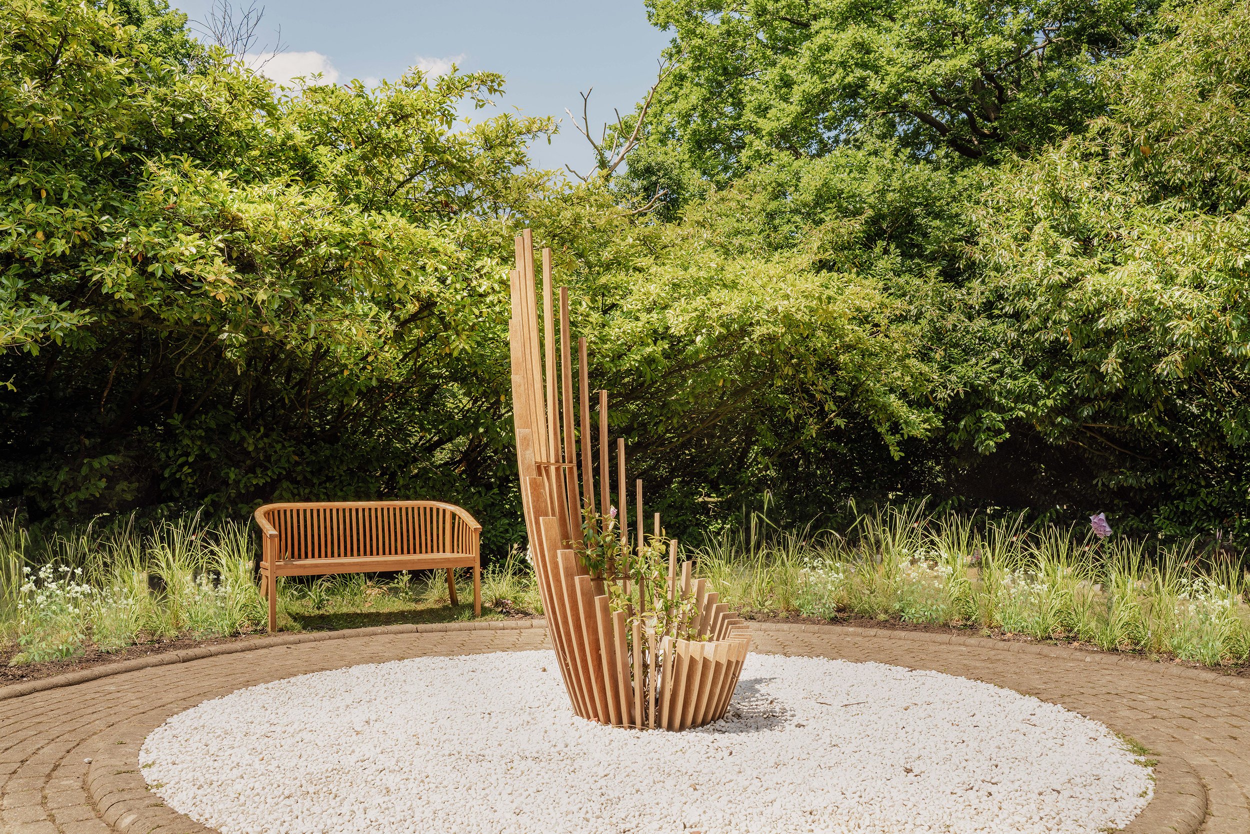 A modern wooden outdoor sculpture with vertical and curved slats, surrounded by a circular bed of white gravel, with a wooden bench and lush green trees and bushes in the background.