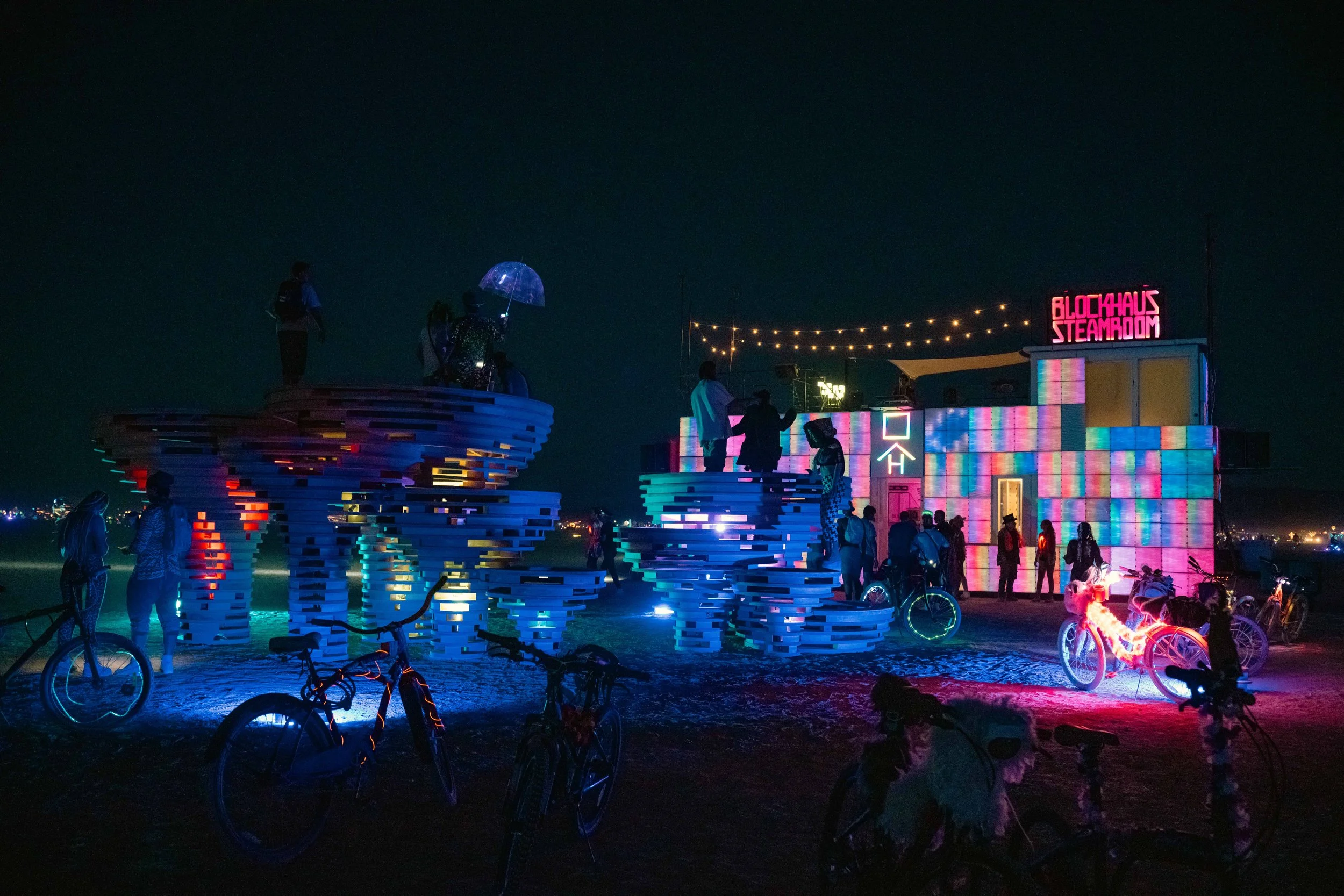 Night scene at Burning Man with illuminated art installations, bicycles, and people gathered around a colorful, glowing structure labeled 'Blockhaus Steamboom' with string lights overhead.
