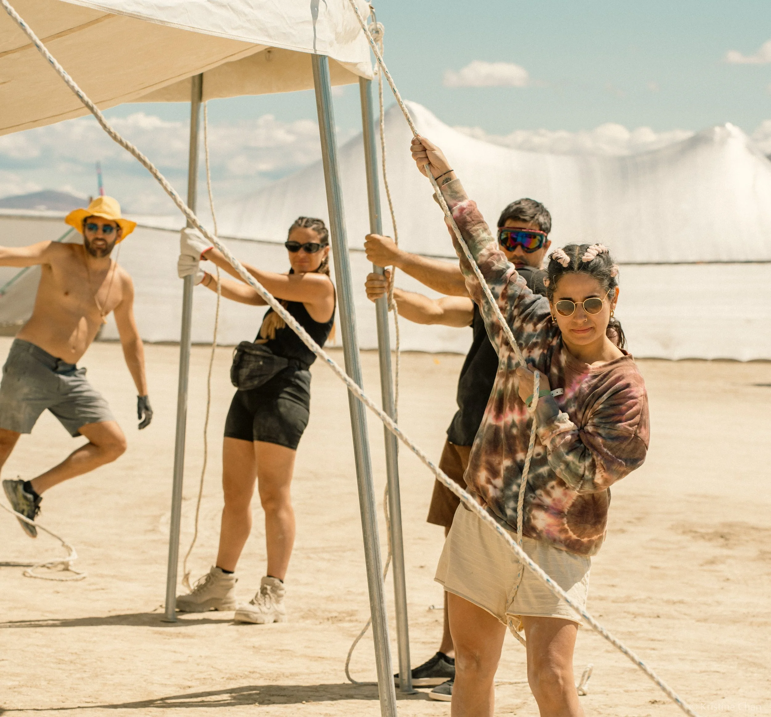 Group of four people holding ropes for a tent or canopy on a sandy desert with salt formations in the background.