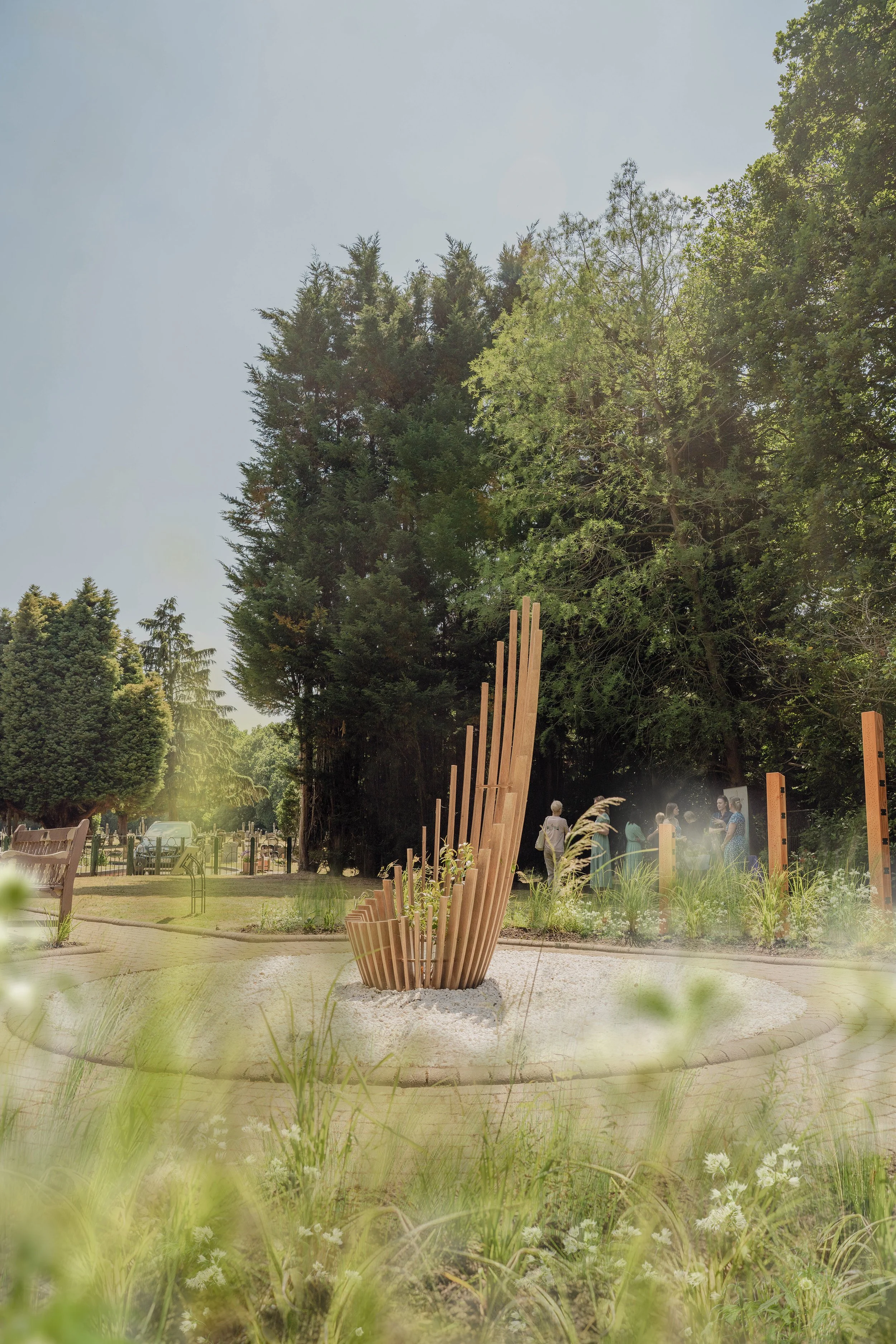 A park with a wooden sculpture in the center, surrounded by green trees, plants, and a few people in the background.