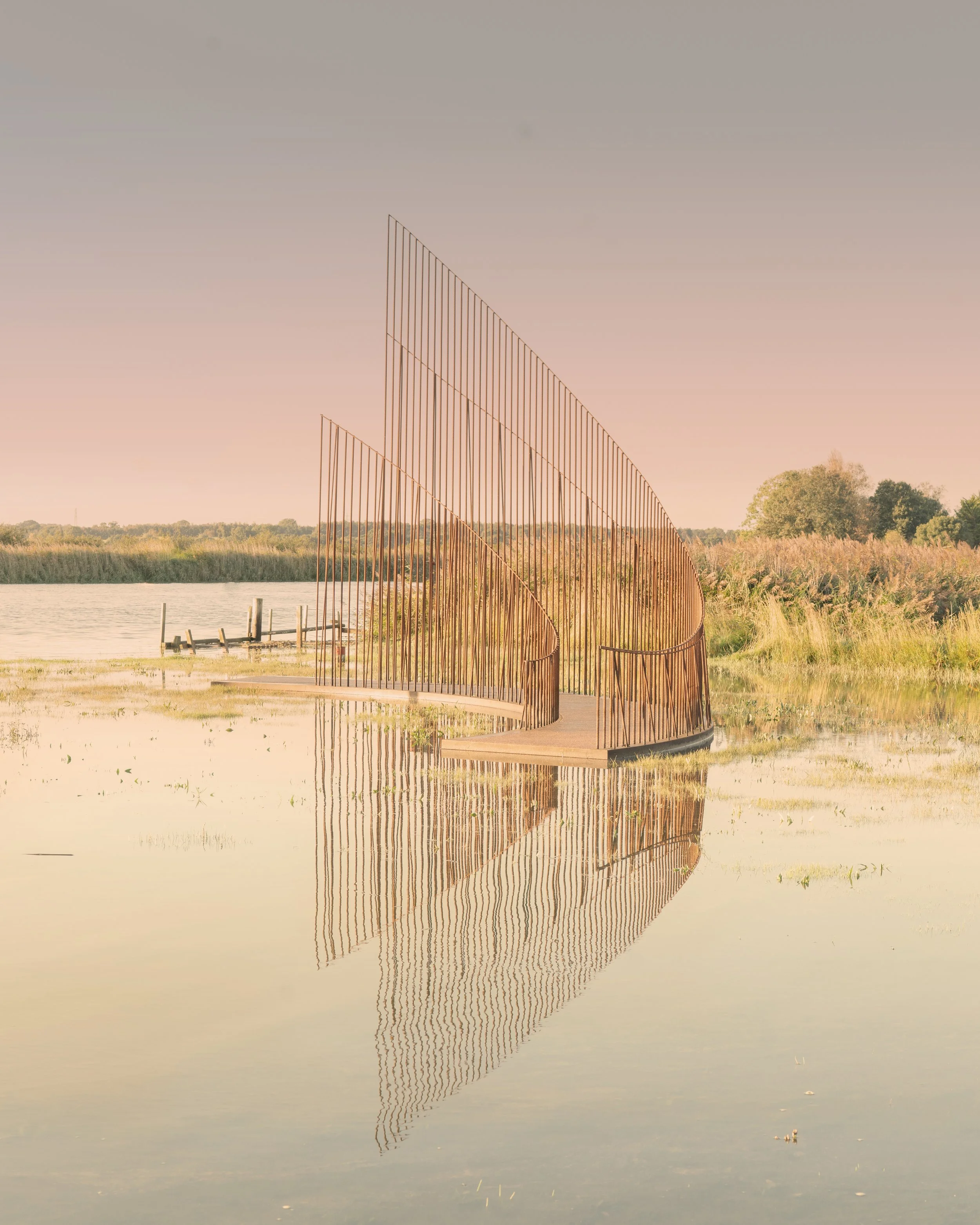 A modern metal sculpture resembling a sailboat reflection in a calm body of water with a grassy shoreline and trees in the background at sunset.