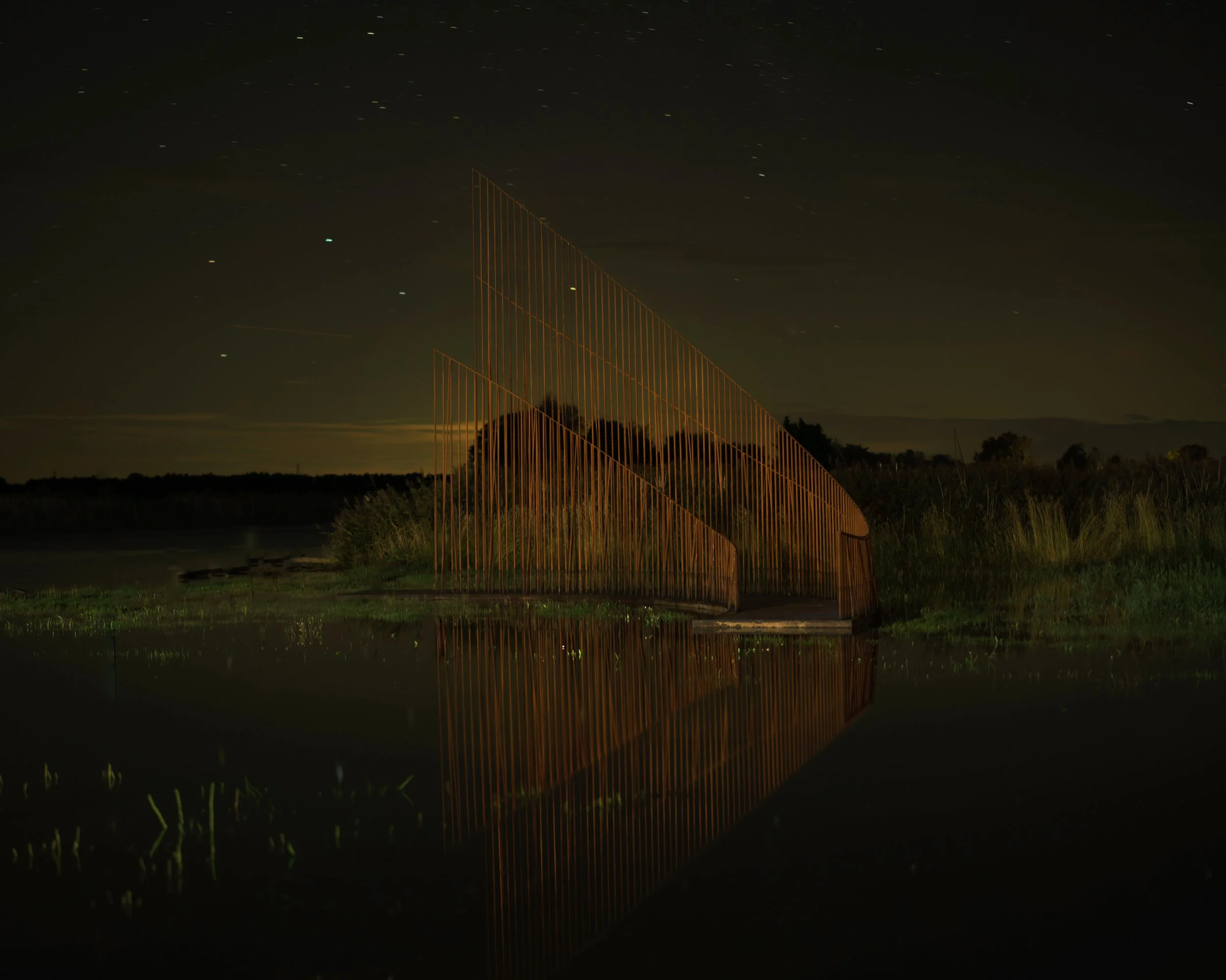 Nighttime view of a large, geometric metal sculpture with colorful light reflections, situated near water and reflecting in it, with starry sky above and grassy terrain in the background.