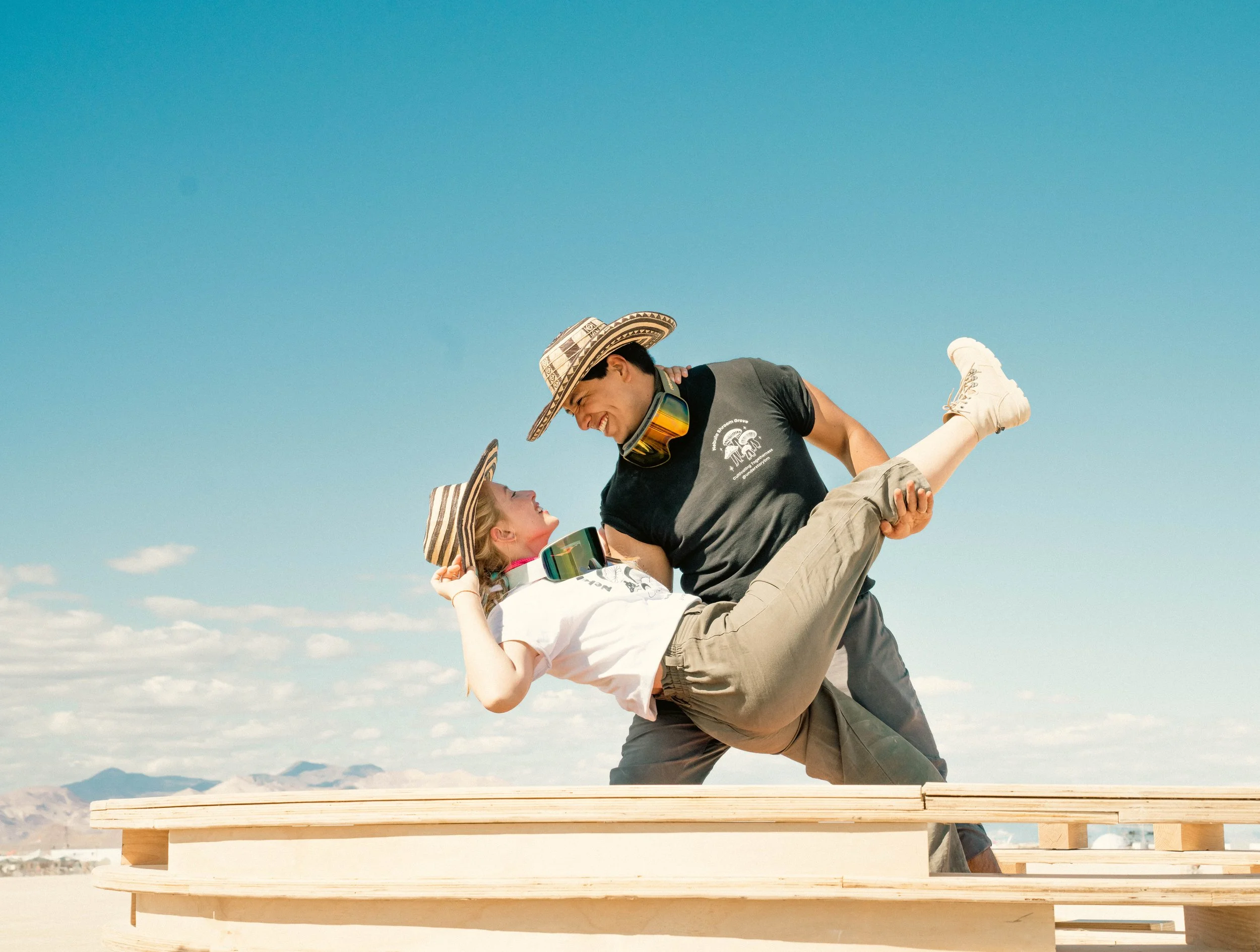 Two people, a man and a woman, wearing hats, are outdoors on a wooden platform with a desert landscape in the background. The man is holding the woman as she leans back, smiling.