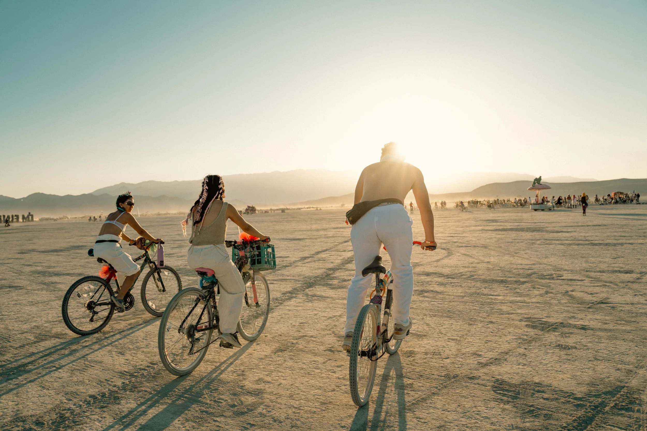 People riding bicycles on a desert at sunset with mountains and a crowd in the background.