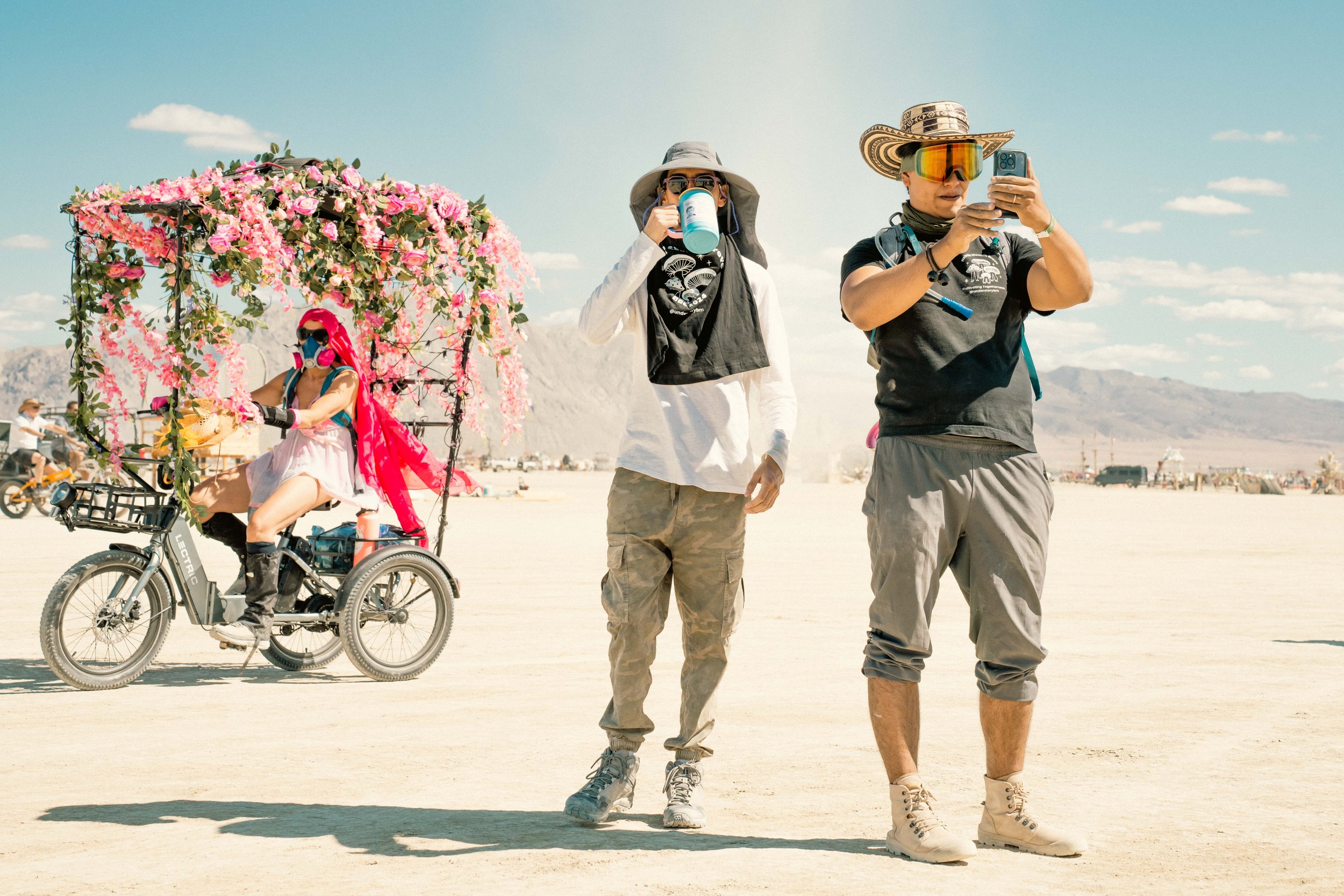 Two people wearing hats and sunglasses taking photos at a desert event, with a woman on a decorated tricycle in the background.