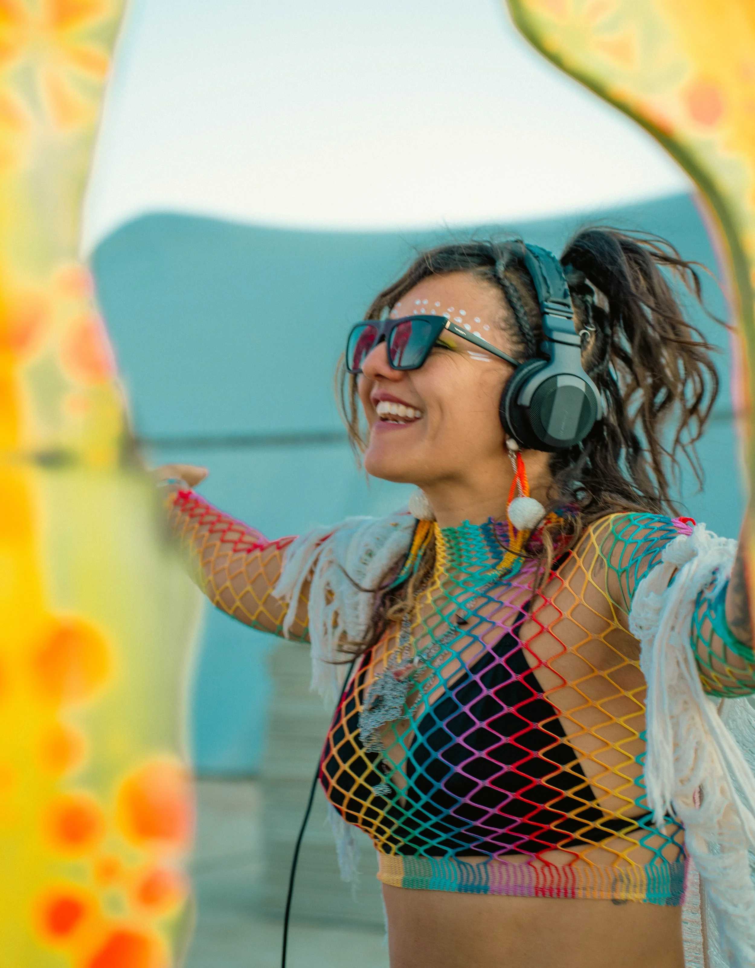 A woman with curly hair wearing sunglasses, headphones, and colorful clothing, smiling with her arms outstretched near a yellow inflatable structure at a beach.