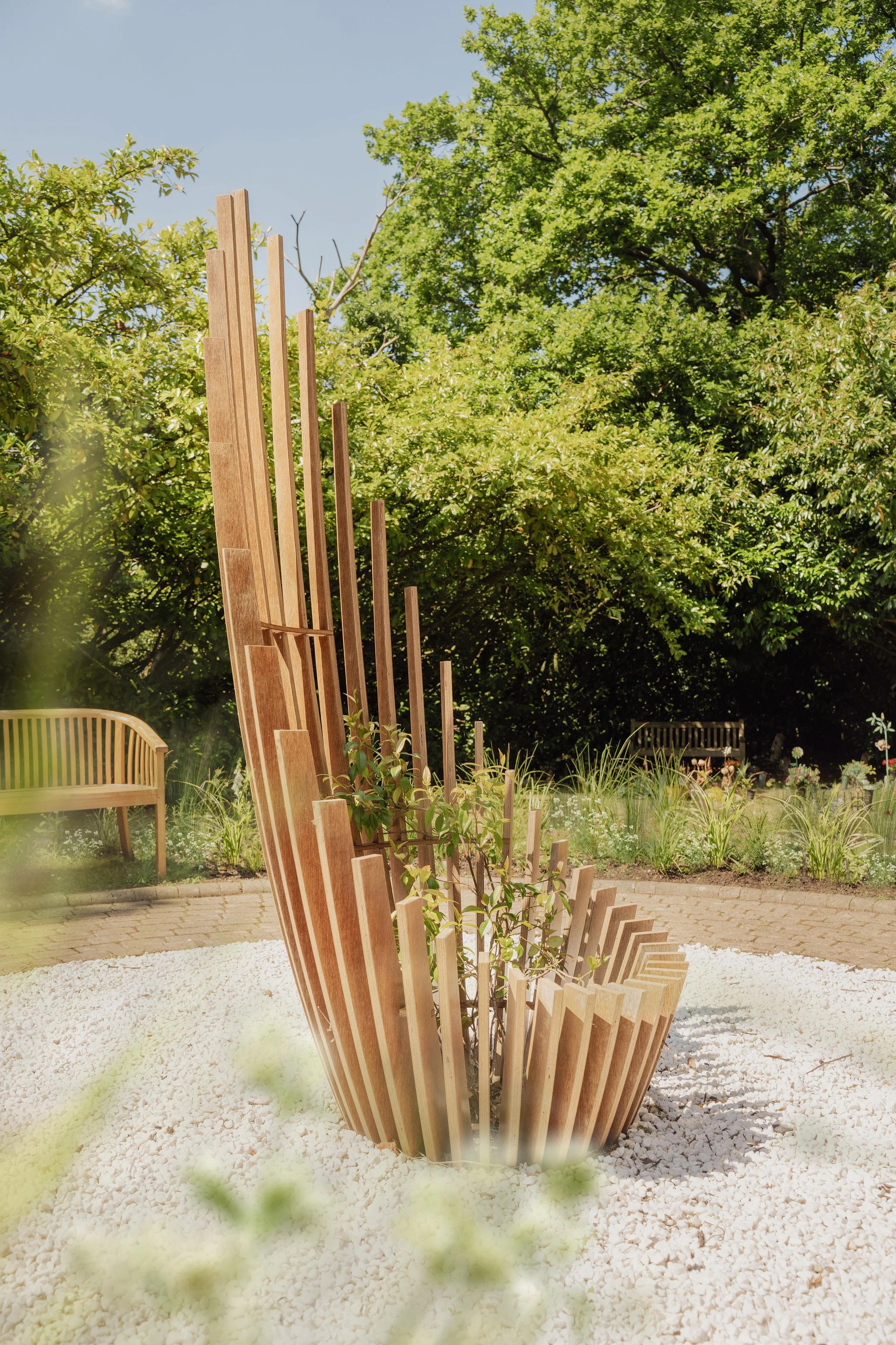 Modern wooden outdoor sculpture with vertical slats surrounded by white gravel, in a garden with green trees and benches.