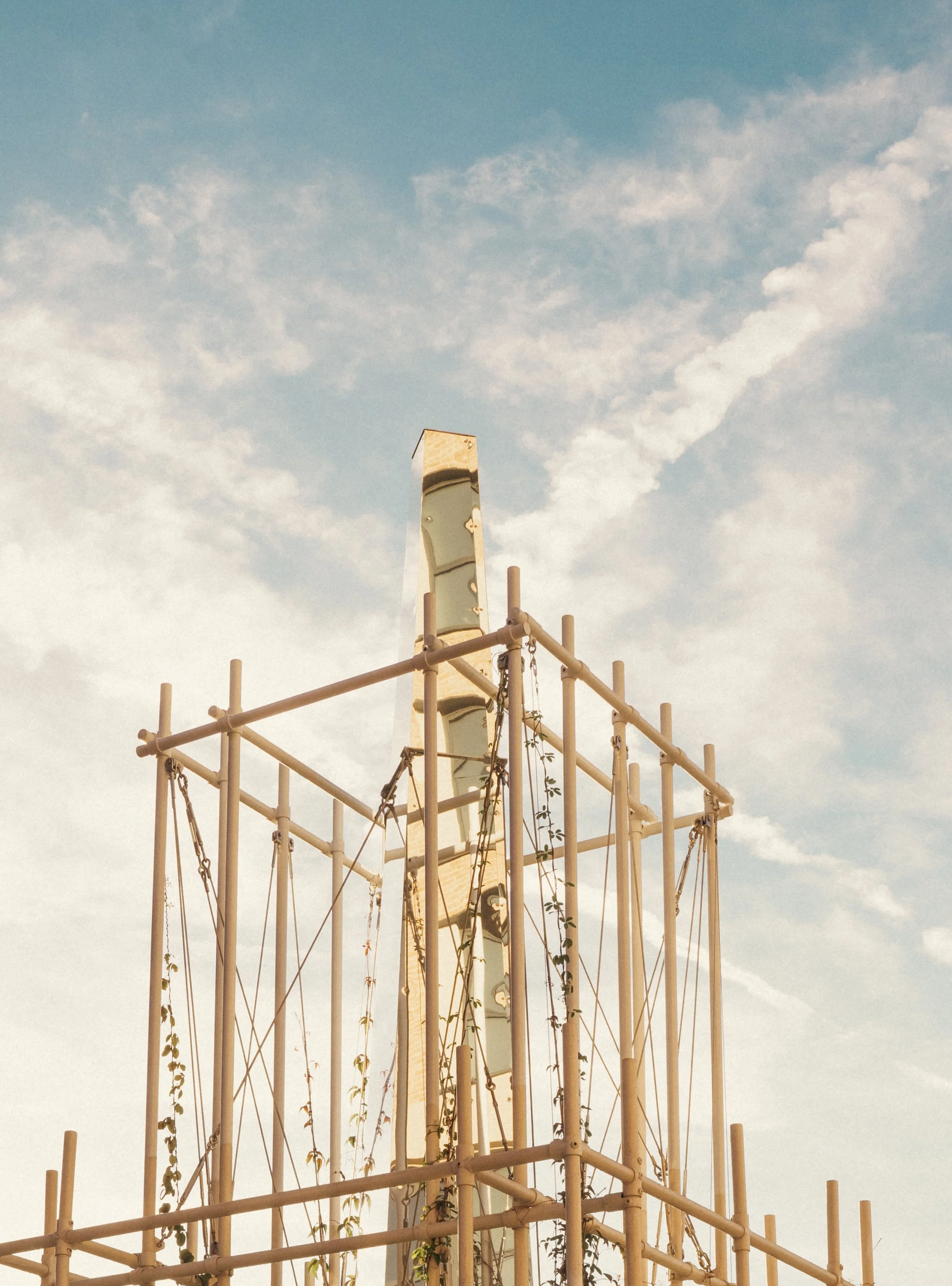 A playground climbing structure with a curved slide or climbing wall, surrounded by a beige metal frame, under a blue sky with scattered clouds.