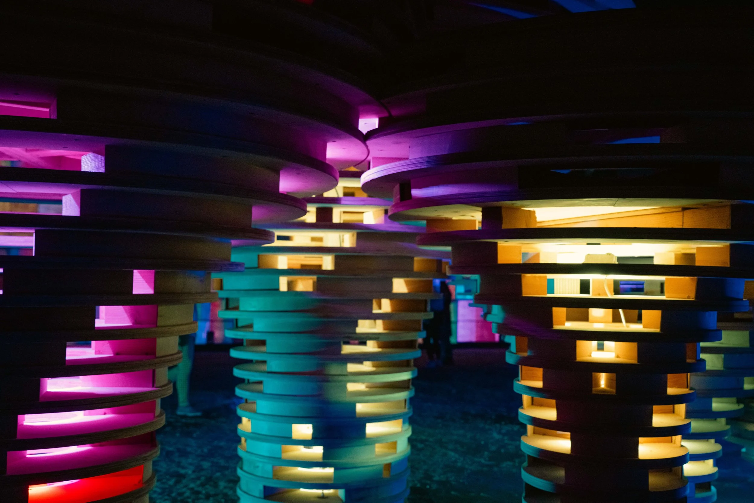 Colorful illuminated wooden structures with stacked circular layers, creating a modern art installation with purple, yellow, green, and pink lighting.