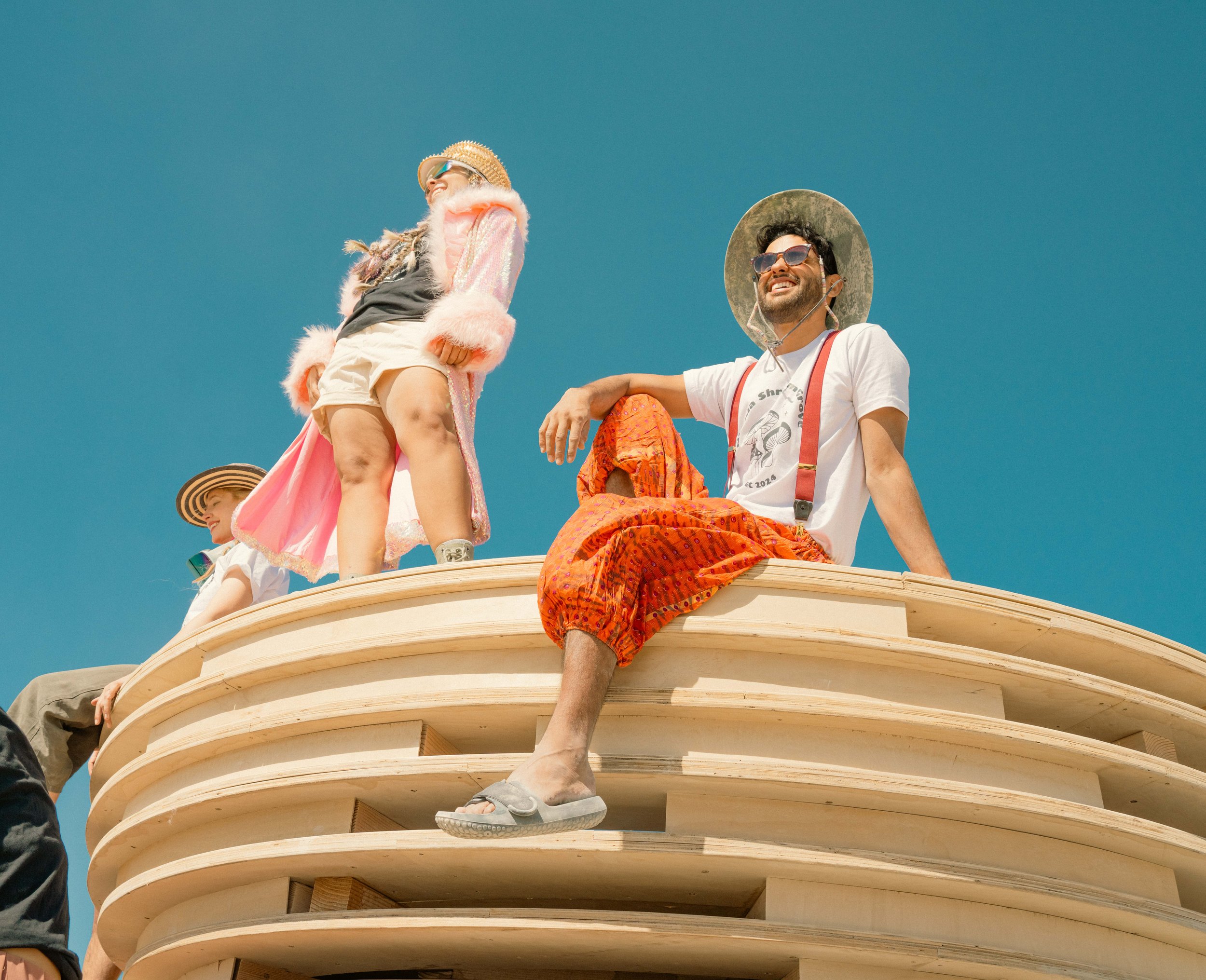 Three people dressed in summer clothing and hats on a rounded wooden structure against a clear blue sky.