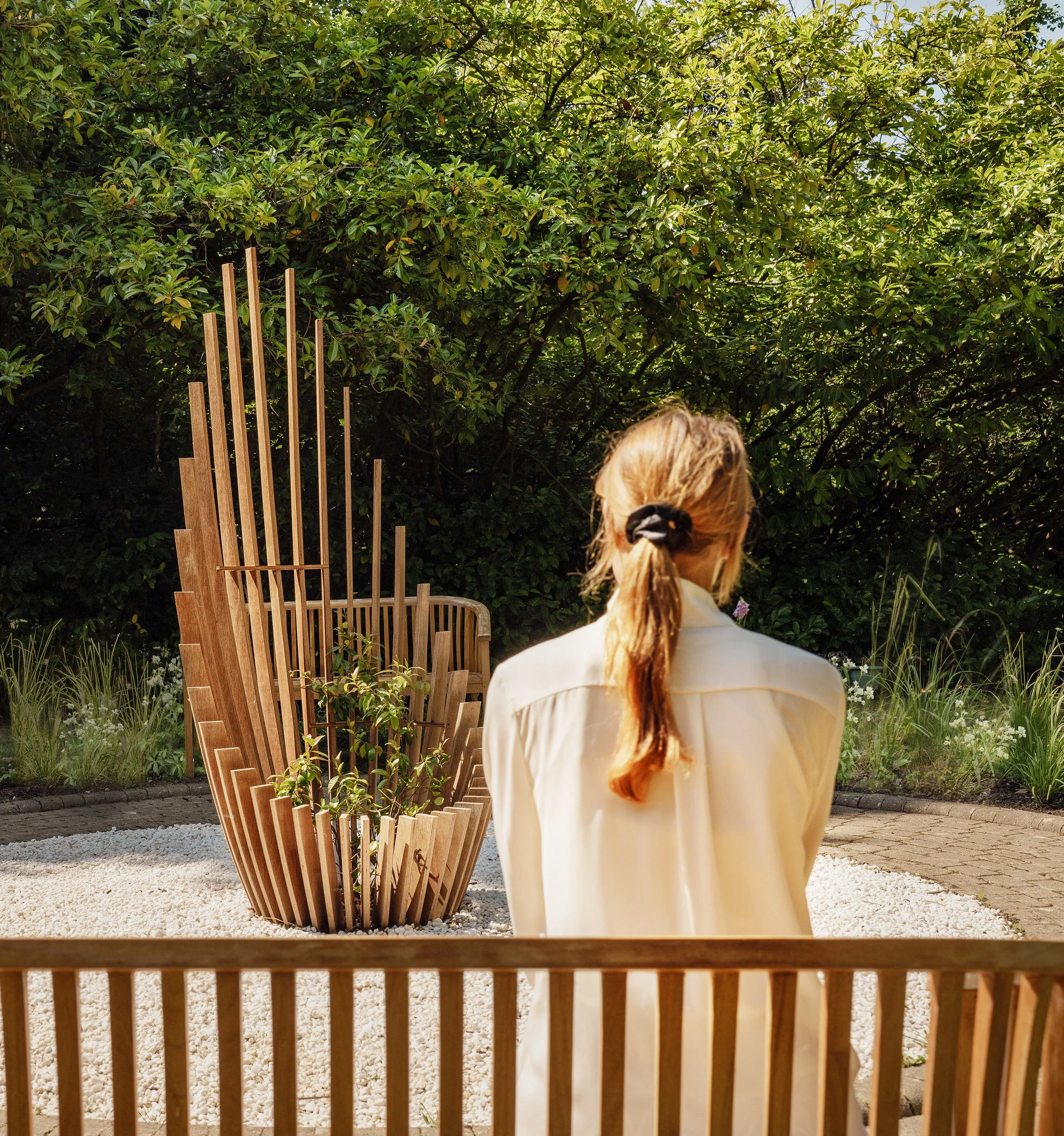 A woman with a ponytail tied with a black scrunchie, dressed in a white shirt, sitting on a wooden bench, overlooks a landscaped garden with a wooden modern sculpture and green foliage.