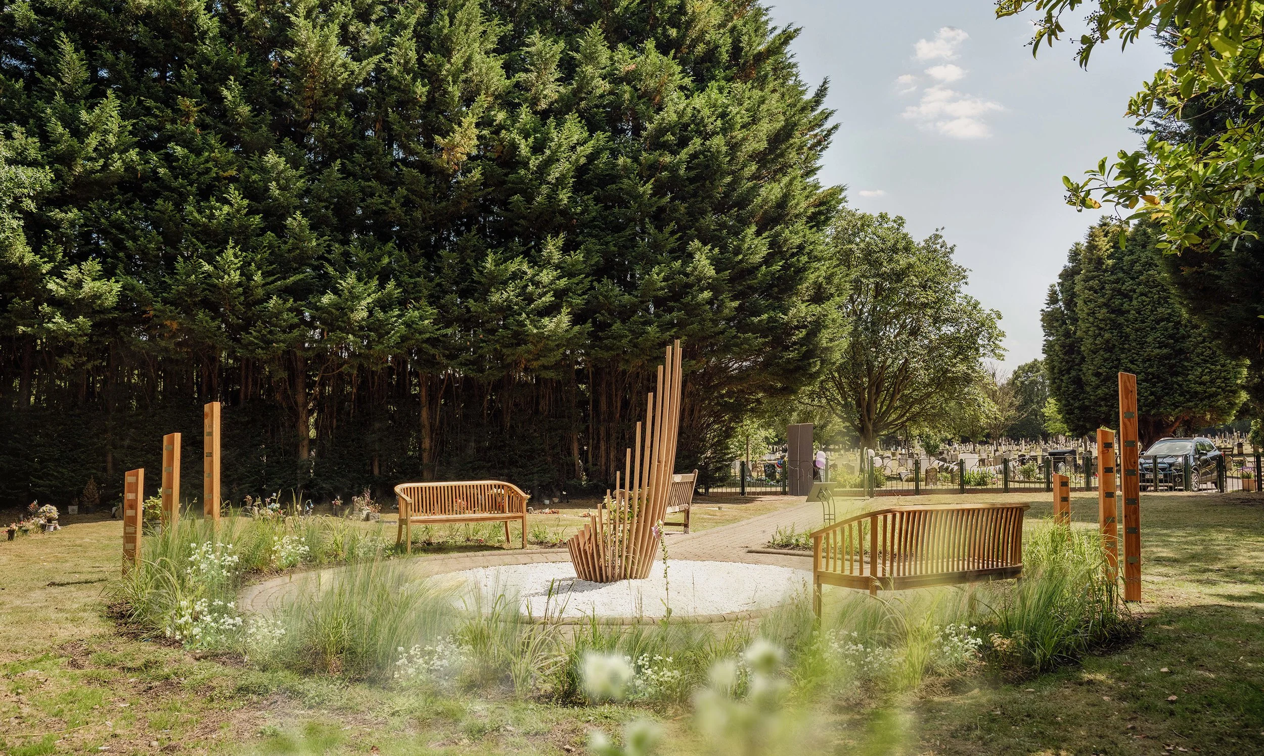 A peaceful park scene with three wooden benches, a gravel pathway, and a modern wooden sculpture in the center. Tall trees and a cemetery with headstones are visible in the background, and the sky is partly cloudy.