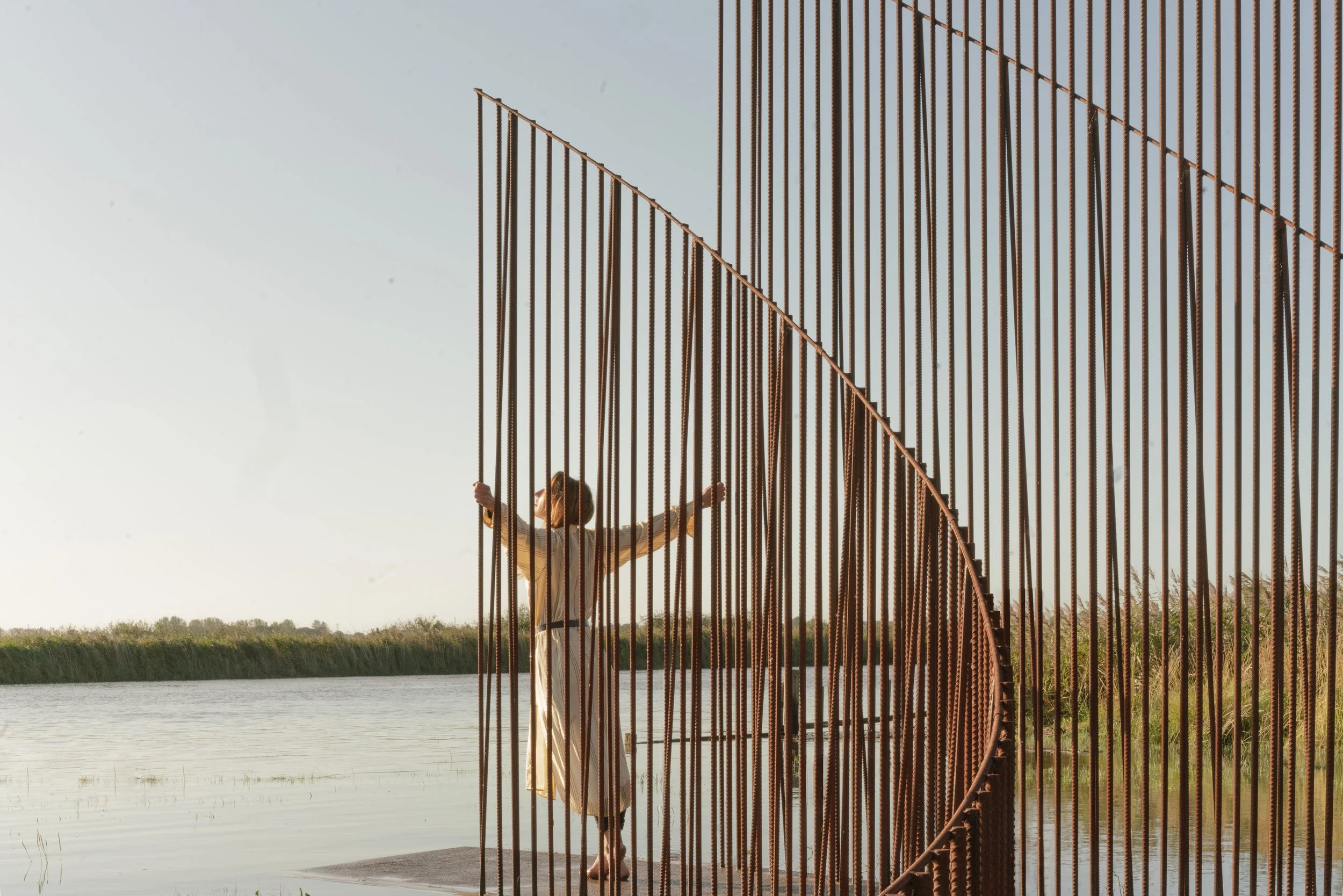 A woman in a beige dress standing inside a large circular metal sculpture near a body of water during sunset.