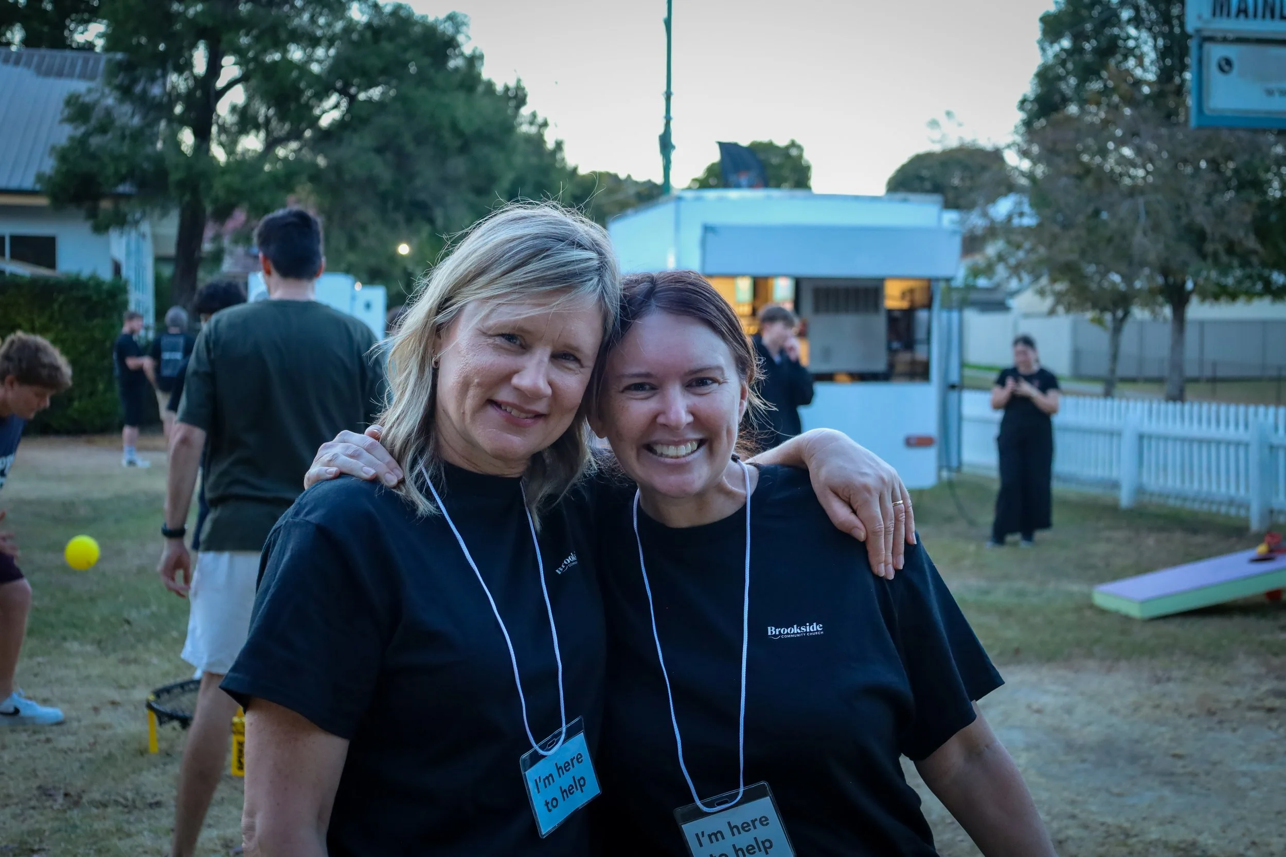 Two smiling women with volunteer badges reading "I'm here to help," hugging outdoors at an event, with a food truck, people, and trees in the background.